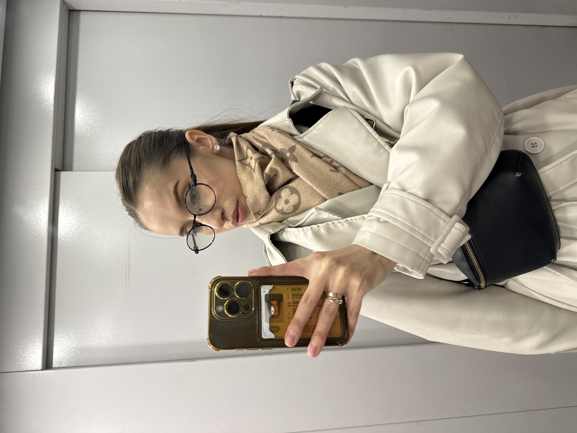 A woman is taking a selfie in what appears to be an elevator with grey walls. She is wearing a light-colored trench coat, a beige scarf with a repeating pattern, and round glasses. She is holding a gold-colored iPhone. A black bag is visible, strapped around her waist, and she is wearing two rings on her right hand. Her hair is pulled back. The environment is utilitarian, suggesting a public or commercial space. The location cannot be determined as Yekaterinburg, Russia from the available image.