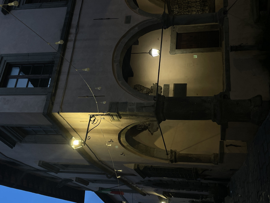 The image depicts the facade of a multi-story building in Castel del Piano, Italy, observed during nighttime or twilight. The structure features light-colored stucco walls and dark stone elements, including two prominent archways at ground level supported by thick stone columns. An antique-style street lamp with a black wrought-iron bracket is mounted on the building's corner, emitting yellow light. A string of approximately twelve bare incandescent light bulbs extends horizontally across the upper portion of the facade. An Italian flag, displaying green, white, and red vertical stripes, is attached to the upper left section of the building. Within the rightmost archway, a dark wooden door is visible, illuminated by a suspended lantern. Decorative stone carvings are present above the archways. The upper floor exhibits dark windows with visible reflections. The foreground consists of a dark cobblestone street or pathway. No individuals, actions, or direct interactions are visible.