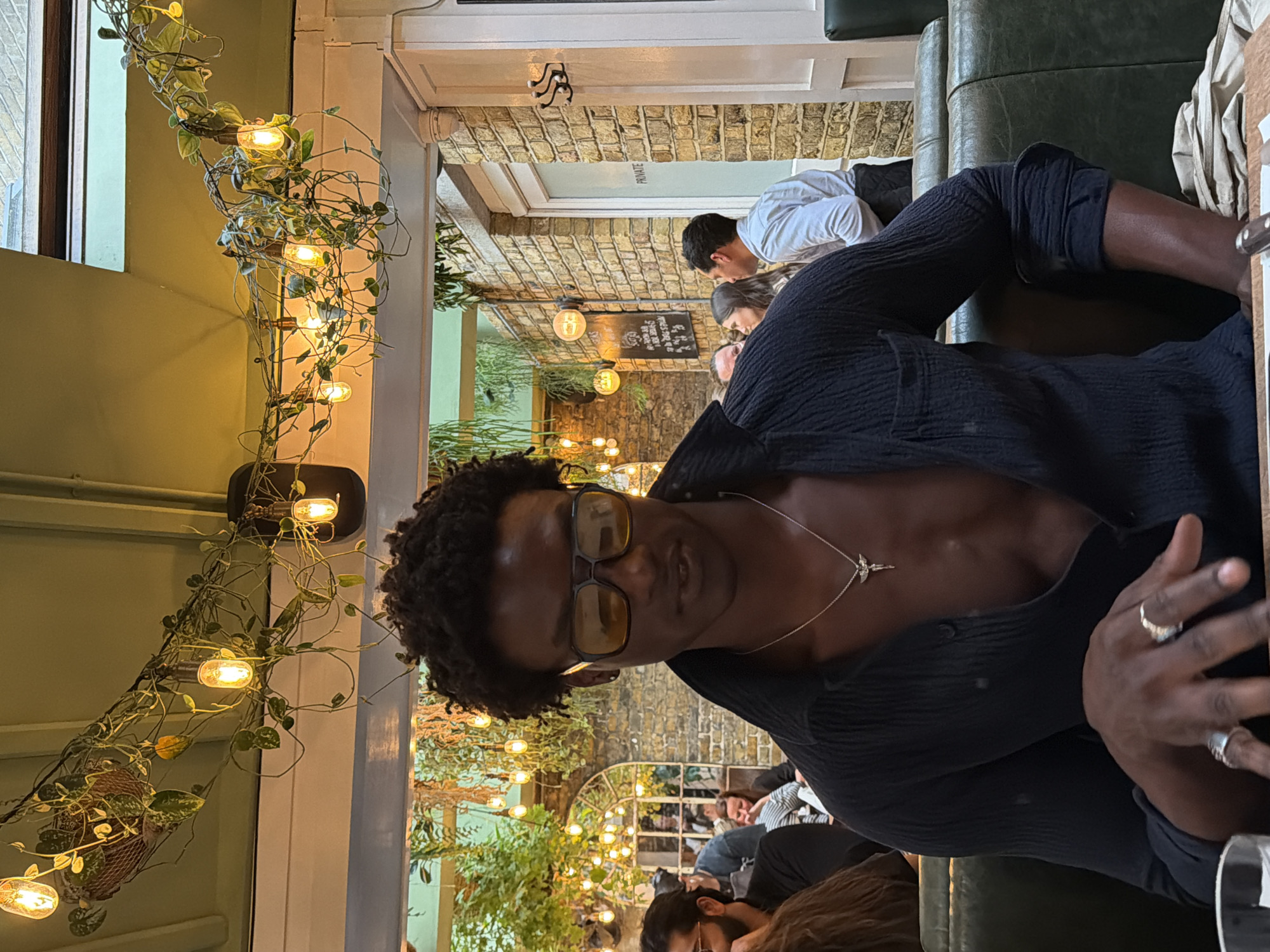 The image, taken in what appears to be a restaurant or cafe in London, United Kingdom, shows a person seated at a table, looking directly at the camera. The main subject has dark, curly hair and is wearing yellow-tinted sunglasses, a black textured shirt unbuttoned to reveal part of their chest, and a silver necklace with a small bird-shaped pendant. Several rings adorn their right hand.
The setting is an indoor space with a warm, ambient glow, suggesting evening or nighttime. To the left of the main subject, a decorative strand of string lights is intricately wrapped around green vines, extending vertically alongside a window. The furniture includes dark green leather upholstery, possibly part of a booth or sofa where the subject is seated.
In the mid-ground and background, other patrons are visible, engaged in conversation. One man in a white shirt is seen interacting with a woman. The interior features exposed brick walls, adding a rustic charm to the establishment. More decorative lights and potted plants are strategically placed throughout the space, enhancing the cozy atmosphere. A doorway or archway is visible in the background, with a frosted glass panel above it that clearly displays the word "PRIVATE." The general activity appears to be casual dining or socializing in a relaxed environment.
