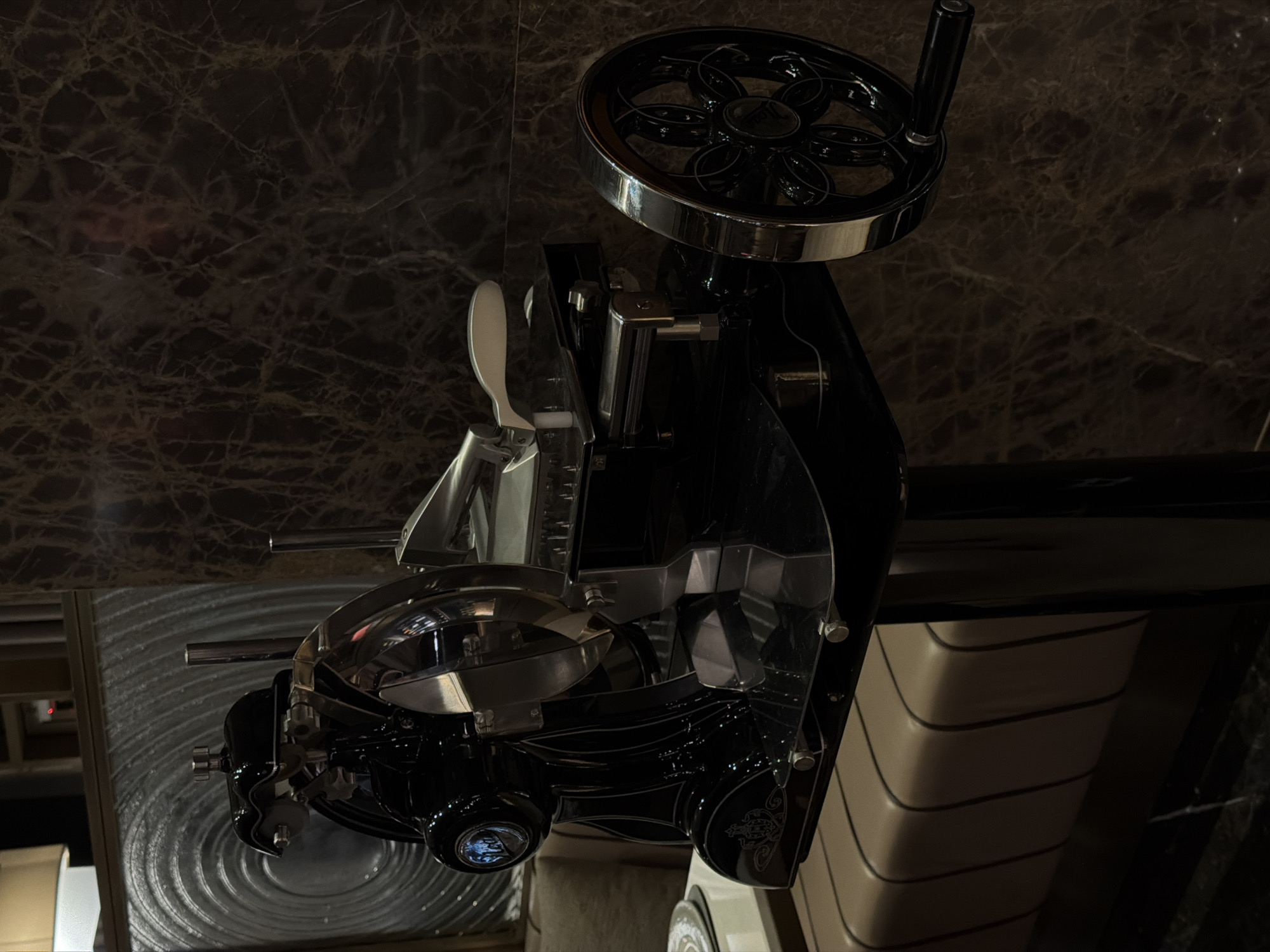 The image displays a sophisticated, retro-style mechanical slicer, possibly for deli meats or charcuterie, set against a dark, richly veined marble background. The slicer is predominantly black with highly polished chrome or stainless steel accents, contributing to its luxurious aesthetic.
The central object is the slicer itself, which features a prominent, spoked hand-crank wheel at the top right, adorned with intricate patterns. Various levers, knobs, and finely crafted metallic components are visible throughout the machine, indicating precision engineering. The base of the slicer exhibits decorative scrollwork or engraving, adding to its vintage charm. To the bottom left, a circular, ridged metallic surface, possibly a serving tray or part of the machine's stand, is visible. Below the right side of the slicer, a segment of furniture or cabinetry with horizontal, concave ridges, in a light beige tone, adds another layer to the setting.
No people are present in the scene, and no activity is taking place; the machine appears to be on display or idle. The lighting is subdued, creating reflections on the metallic surfaces and emphasizing the machine's sleek design. The background's dark marble suggests an elegant indoor setting, such as a high-end delicatessen, restaurant, or a luxury kitchen display in Budva, Montenegro. A faint, stylized logo, possibly a letter or symbol, is discernible on a circular knob at the front of the slicer, but no legible text is visible elsewhere.
