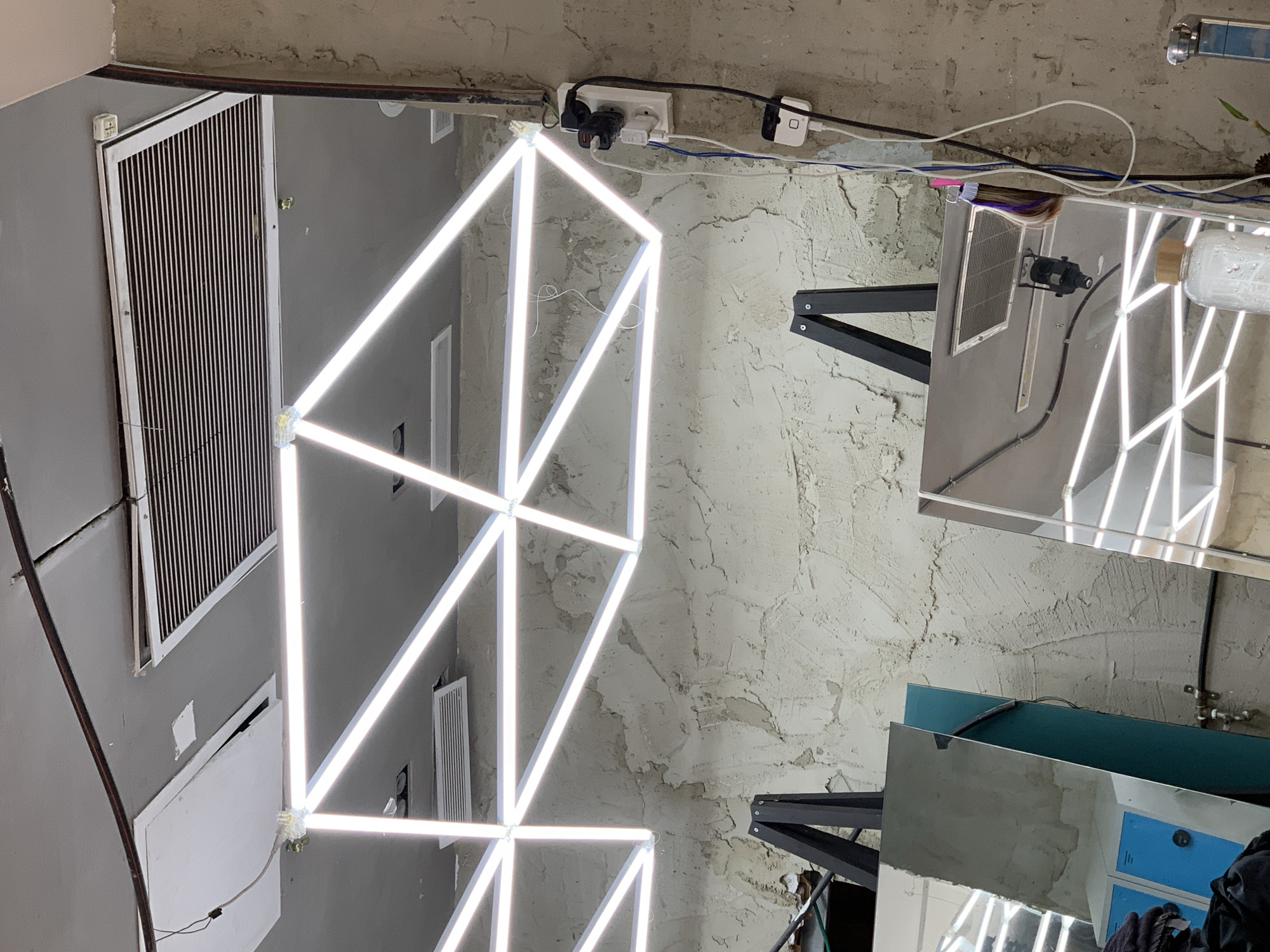 The image depicts the interior of a building, possibly a business, with a focus on the ceiling and upper walls. The location is unknown, and the claim of being in Tel-Aviv, Israel, cannot be verified.
The ceiling has a rough, textured surface. A large, geometric light fixture consisting of connected, illuminated bars dominates the scene. Electrical outlets and wiring are visible along the upper wall near the ceiling.
Parts of interior walls are visible. Also visible are reflective surfaces, possibly mirrors, and the top of what appears to be a cabinet or furniture piece with a blue interior. A person wearing black clothes is visible. No interactions between individuals are evident. The scene appears static.