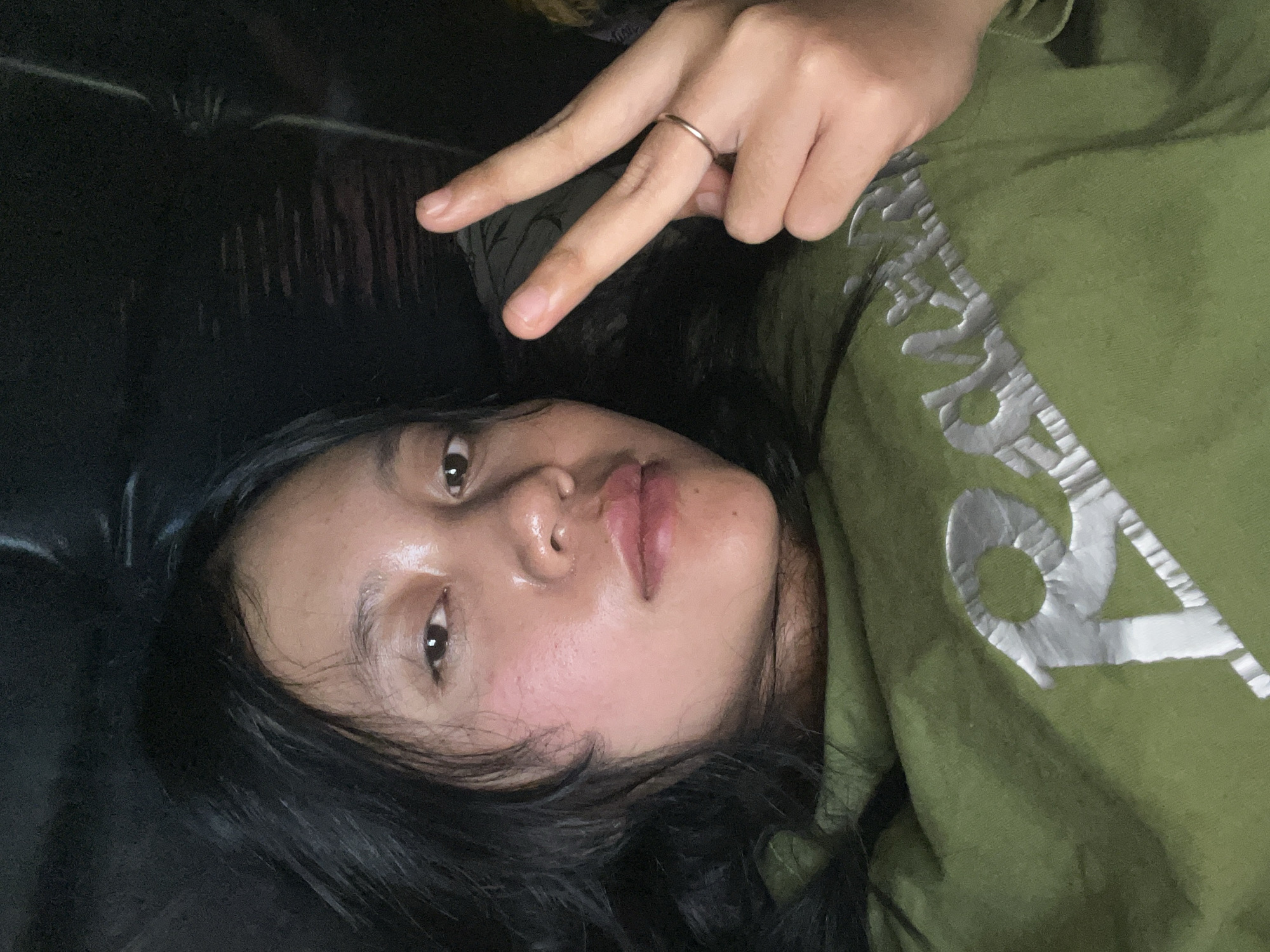 A young woman is shown in a close-up, selfie-style photograph, lying down and looking directly at the camera. Her long, dark hair frames her face, and she has fair skin with a slightly flushed complexion. She is wearing a green hooded sweatshirt with a metallic silver graphic design on the front. Her right hand is raised, with two fingers extended in a peace sign. A thin, gold ring is visible on her ring finger. The background is dark and indistinct, suggesting an indoor setting. The image is well-lit, focusing on the woman's face and upper body. There are no visible elements that place this image in Jambearum, Indonesia; the description is limited to the visual information present.