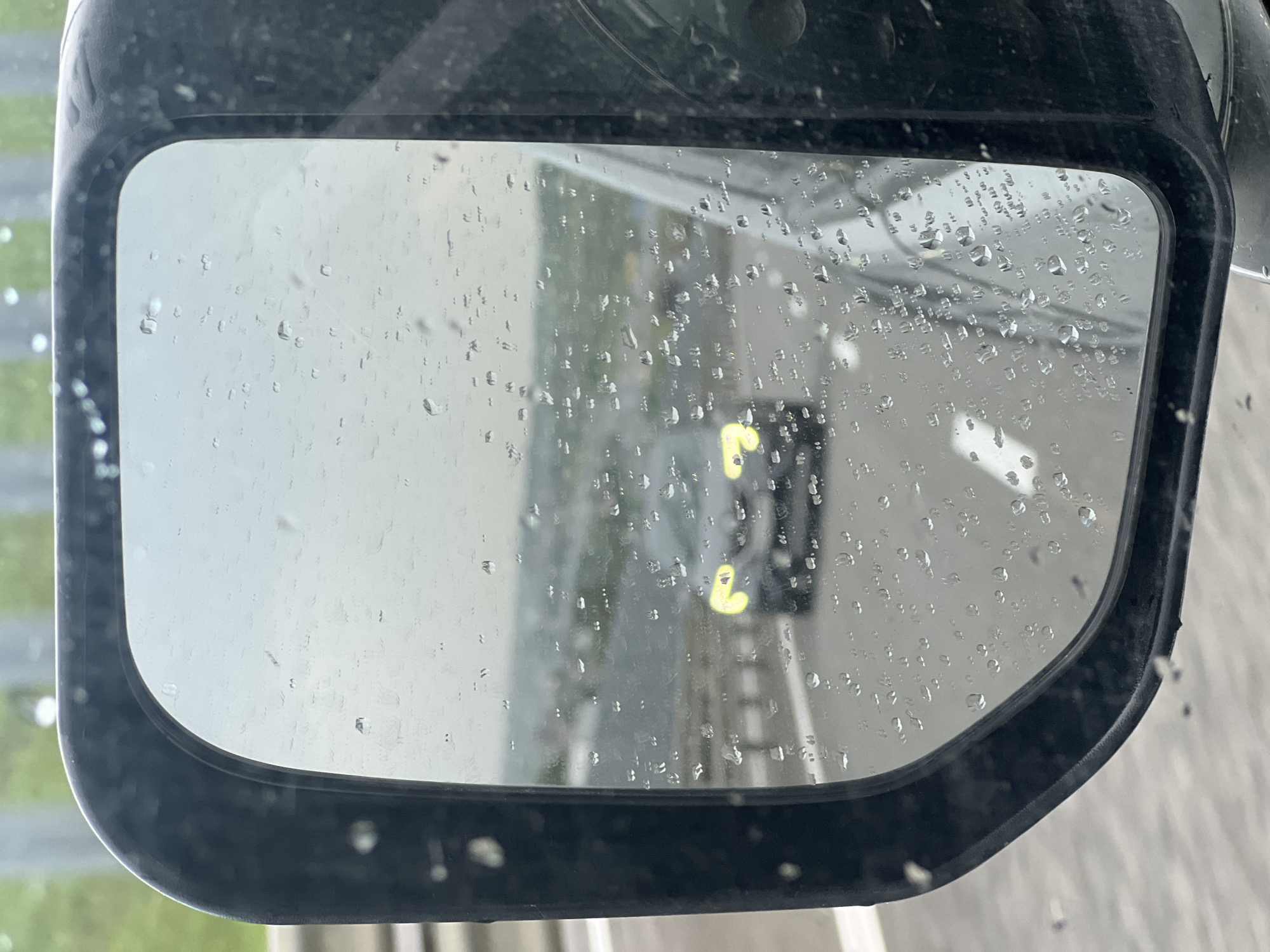 A rain-streaked side-view mirror of a vehicle reflects a fire engine driving behind. The mirror itself is black. Raindrops cover the surface of the mirror, obscuring the clarity of the reflection. The fire engine appears to have lights flashing. The environment outside the mirror shows a wet road surface and indistinct surroundings. The location is unknown.