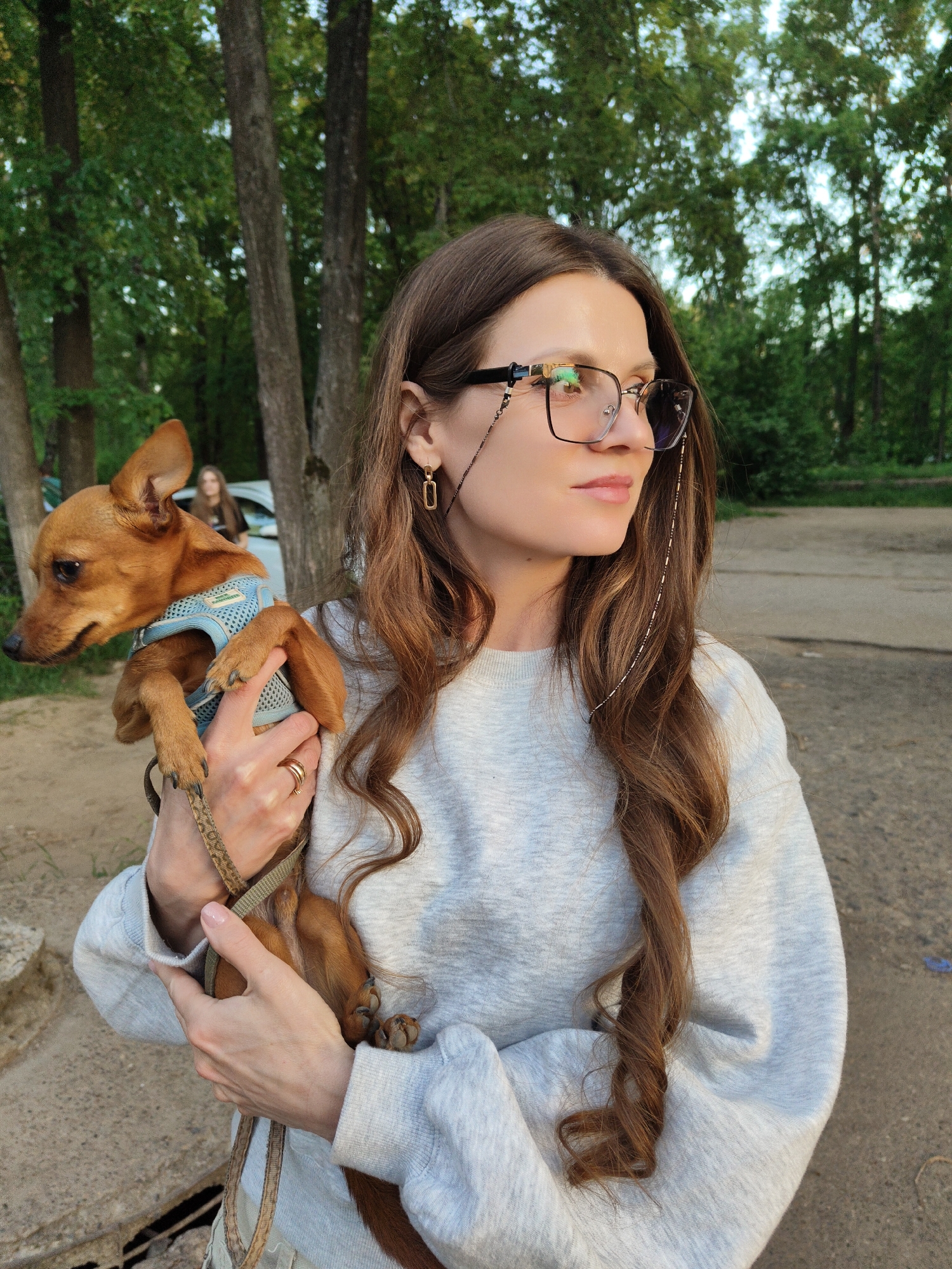 A medium shot depicts a woman holding a small brown dog in her arms. The woman has long, wavy brown hair and wears glasses with a chain. She is wearing a light grey sweatshirt. She gazes off to the right of the frame. The dog is held with a light blue harness, and its leash is loosely held in the woman's hand. The background consists of trees and a dirt path, suggesting a park setting in the city of Unknown, Unknown. In the distance, there is a person and a vehicle.