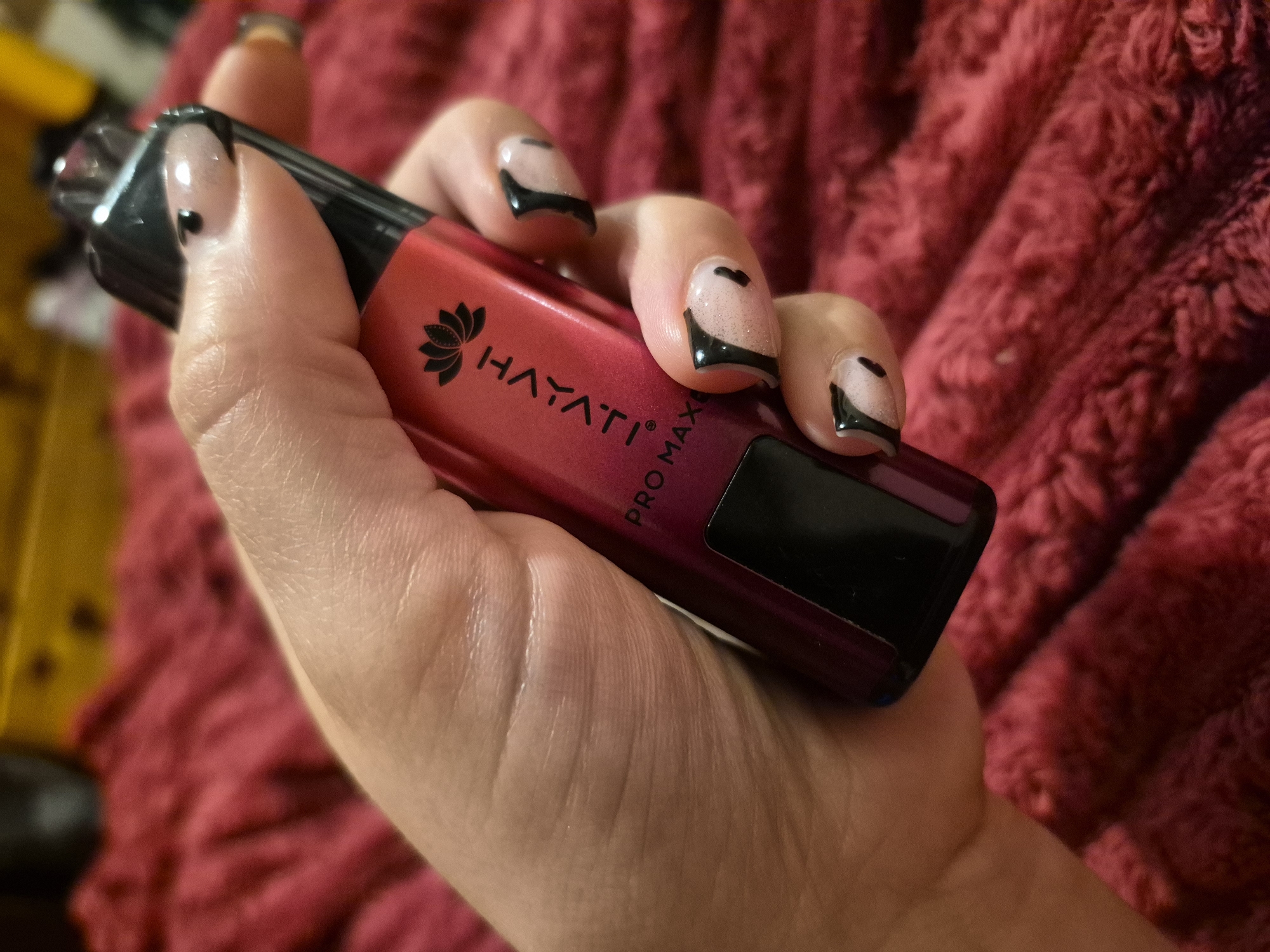 A close-up, eye-level shot shows a person's hand holding a red and black disposable vape. The vape has the brand name "HAYATI" and "PRO MAX" printed on it in white text, with a black lotus flower logo above the brand name. The person has black and nude-colored nail art. The background is a blurred, textured red surface, possibly a blanket or rug. The location is not specified in the image.