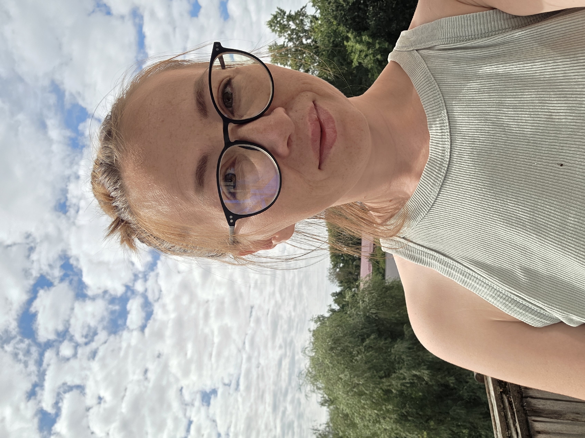 A close-up, selfie-style portrait captures a woman with light brown hair pulled up in a bun, wearing black-rimmed glasses and a light green ribbed tank top. She is positioned against a backdrop of a cloudy blue sky and lush green trees, suggesting an outdoor setting. The perspective is from the chest up, with the woman looking directly at the camera. The lighting is bright, indicating daytime. The scene appears to be in a residential area, possibly a balcony or rooftop, given the wooden railing visible on the left side of the frame. This image was taken on July 16, 2023, in Chaikovsky, Russia.