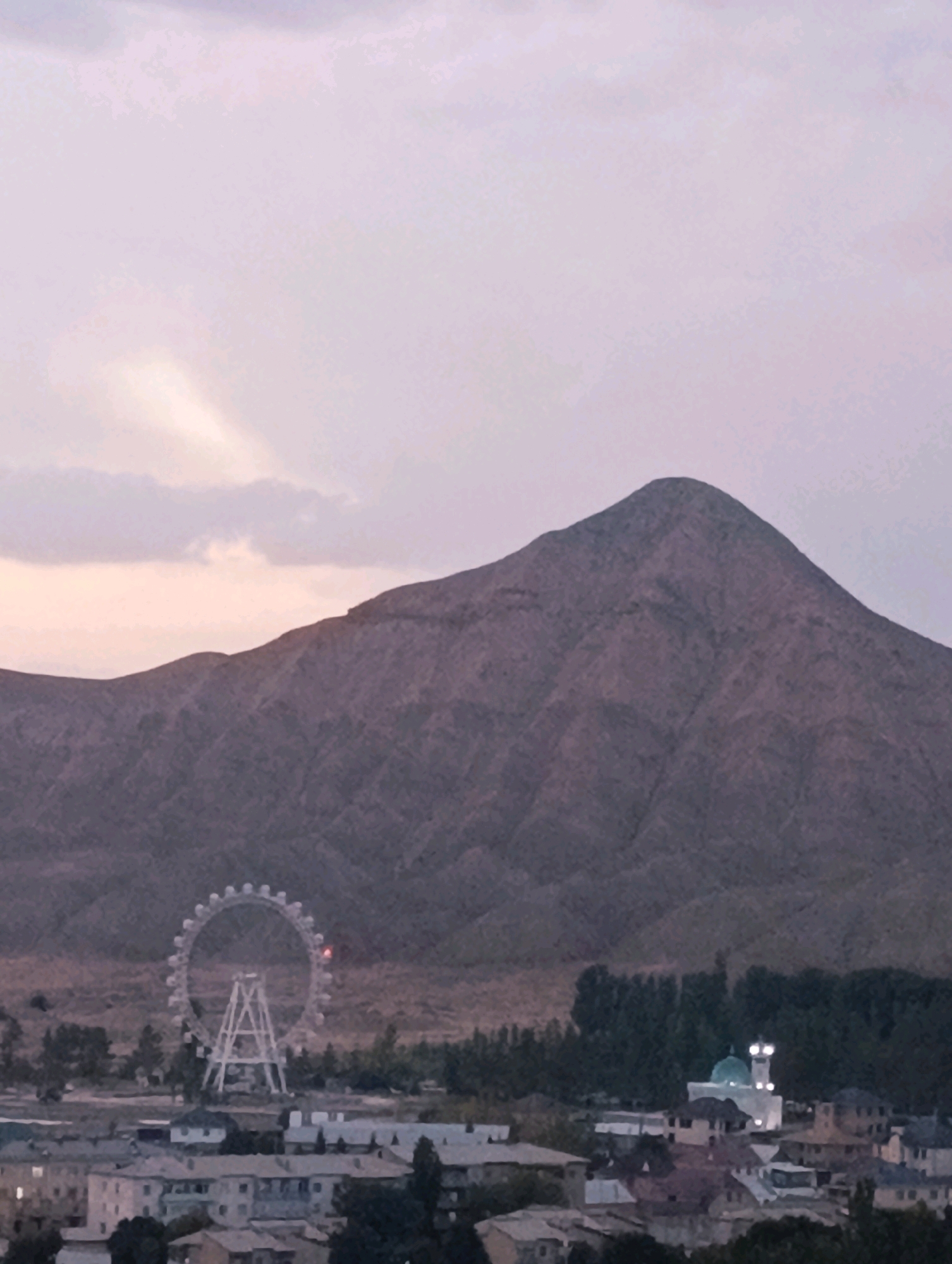 The image depicts a panoramic view of Naryn City, Kyrgyzstan, at dusk. A prominent, rugged mountain dominates the background, its slopes textured and colored in shades of brown and purple. In the middle ground, a Ferris wheel with white spokes and cabins stands out against a dry, scrubby landscape. To the right of the Ferris wheel, a cluster of trees separates it from a white mosque with a turquoise dome, illuminated by lights, and several other buildings in the foreground. The foreground consists of a dense arrangement of residential buildings with varying rooflines and architectural styles, suggesting an urban environment. The sky is a soft, pale lavender with wispy clouds, and a faint light beam pierces through them on the left side. The overall atmosphere is calm and serene, indicative of early evening. There are no people visible in the image.