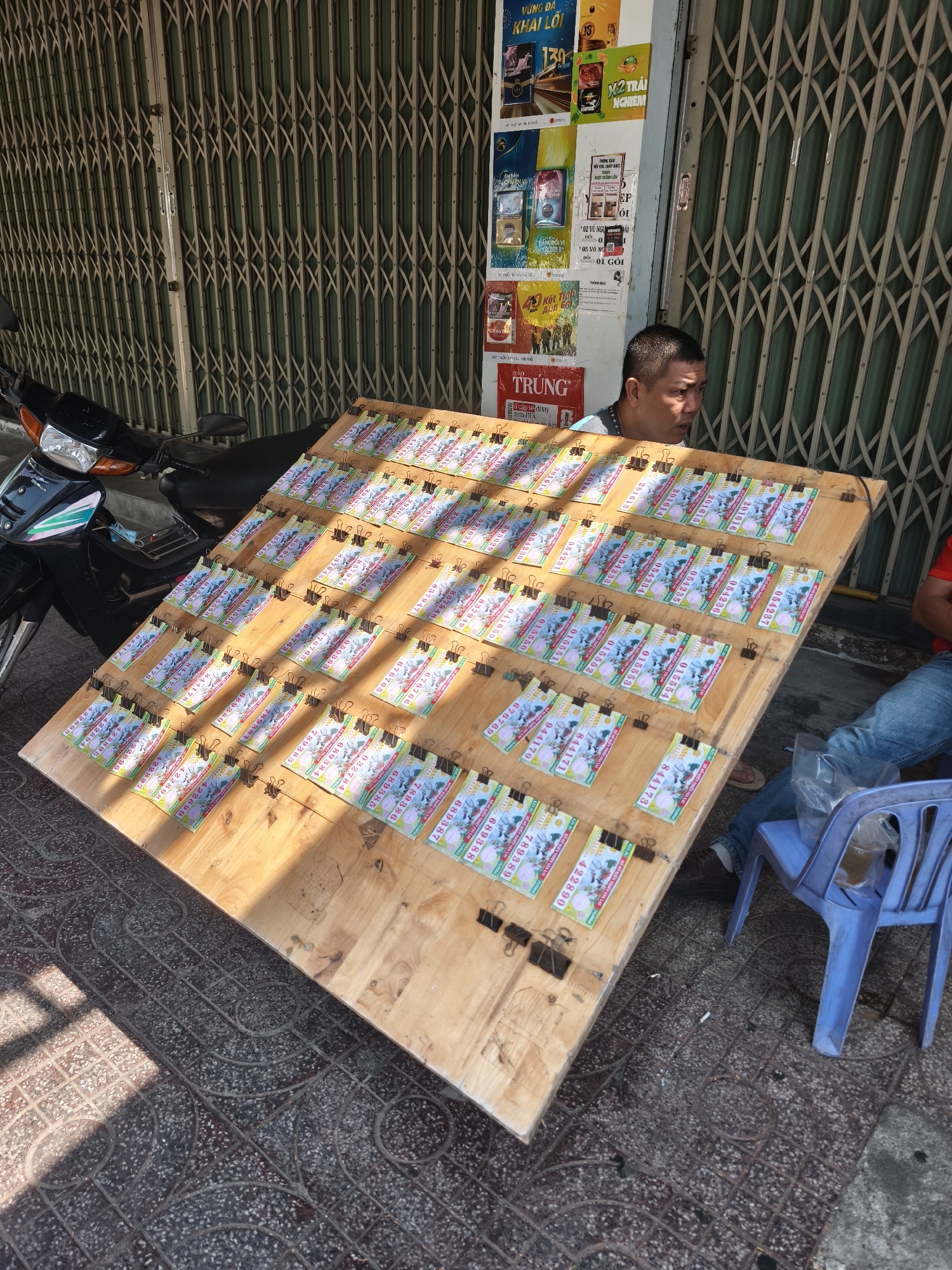 A man is selling lottery tickets laid out on a wooden board, positioned on a tiled sidewalk in front of a closed metal gate. He is seated and looking to his right, with a motorcycle parked behind him to the left. Another person, wearing blue jeans, is seated on a blue plastic chair to the right, with a plastic bag on the ground next to them. The lottery tickets are colorful and feature various numbers and graphics. The scene is illuminated by sunlight, casting shadows on the ground and the lottery tickets. Posters with text and images are affixed to the wall behind the man, suggesting a commercial setting. The overall environment appears to be an outdoor urban space in Bac Nha Trang Ward, Vietnam.