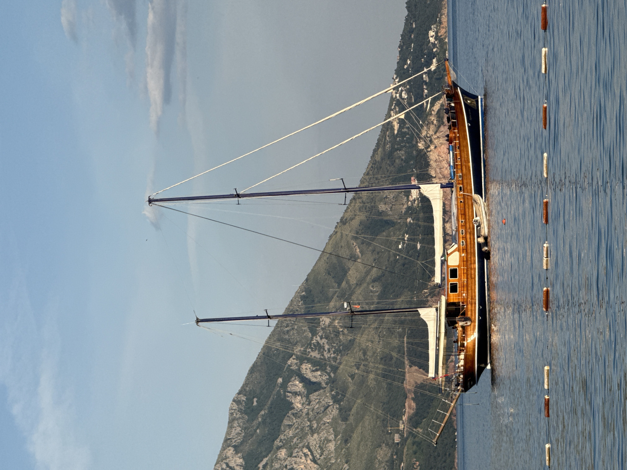 A horizontal, medium-shot photograph shows a sailboat on the water near land. The sky is mostly light blue, fading to a light gray towards the top right corner. There are some streaks of white clouds in the top left corner. On the left side of the image is a tall mountain range, colored green and brown, with a town built into the side of the mountain. A tall sailboat is positioned at the center and right of the image, sitting on calm, blue water. The sailboat is made of dark wood and has two tall masts. The masts have white ropes attached to them. On the right side of the sailboat, there is a line of white and brown buoys that appear to separate the swimming area from the boat harbor. The location is the city of Budva, Montenegro, likely the harbor or near the coast.