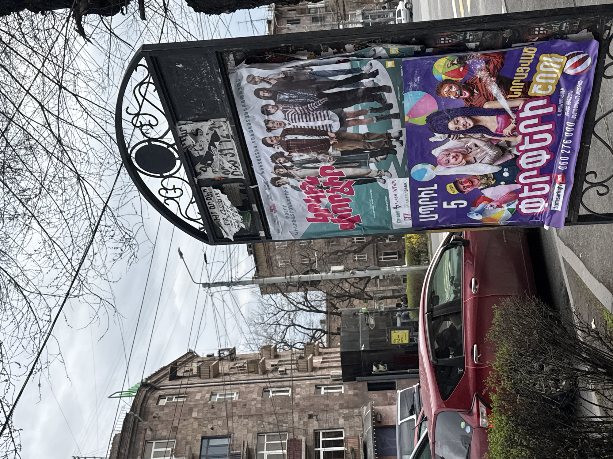 The image shows a street scene, likely in Unknown, Unknown, indicated by the Armenian script on the posters. A black, ornate metal structure holds two posters. The left poster features a group of people. The right poster features clown-like figures. A red car is parked to the right of the metal structure, partially obscured by a bush. A stone building is in the background with many power lines visible in the sky. The sky is overcast.