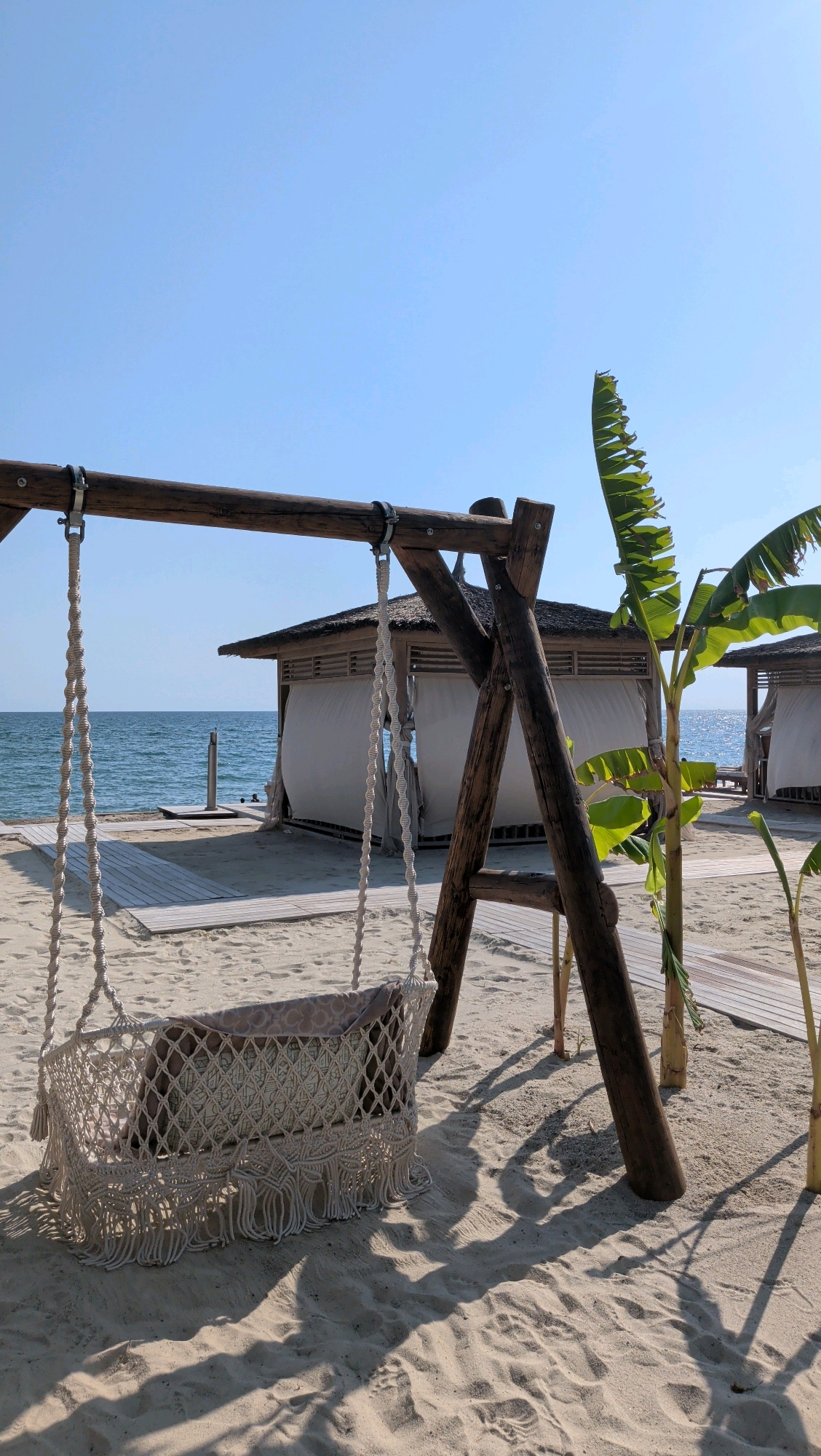 The image depicts a serene beach scene in Serik, Turkey. In the foreground, a wooden swing with a macrame basket seat is set on a sandy beach. The swing is adorned with a patterned cushion. To the right of the swing, a banana tree stands, its large green leaves reaching towards the clear blue sky. Behind the swing, several cabanas with thatched roofs and white curtains are visible, suggesting a resort or private beach area. The ocean stretches out in the background, its surface calm and reflecting the bright daylight. Wooden pathways are laid out on the sand, leading towards the cabanas and the water. The overall environment is tranquil and sun-drenched, typical of a coastal vacation spot.