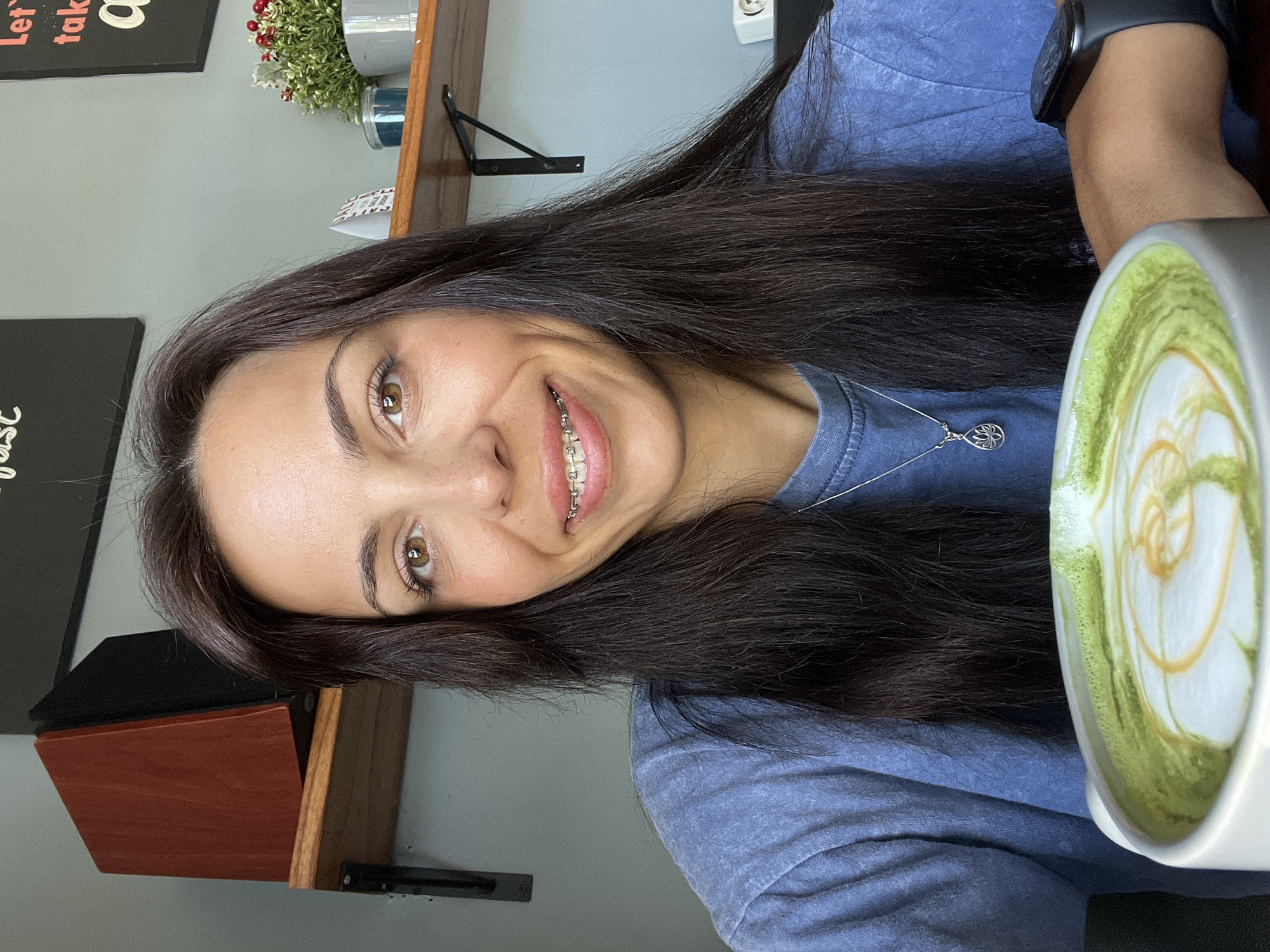 A woman with long, dark hair smiles at the camera, revealing braces. She wears a blue shirt and a delicate silver necklace. To her right, a white mug filled with a green and cream-colored beverage, likely a latte with latte art, is visible. The woman wears a black watch on her left wrist. Behind her is a neutral-colored wall. A small wooden shelf holds greenery in pots and framed pictures, suggesting an indoor setting. The location in the city of Kuta Selatan, Indonesia, is not explicitly evident in the image and would require additional information or landmarks to confirm.
