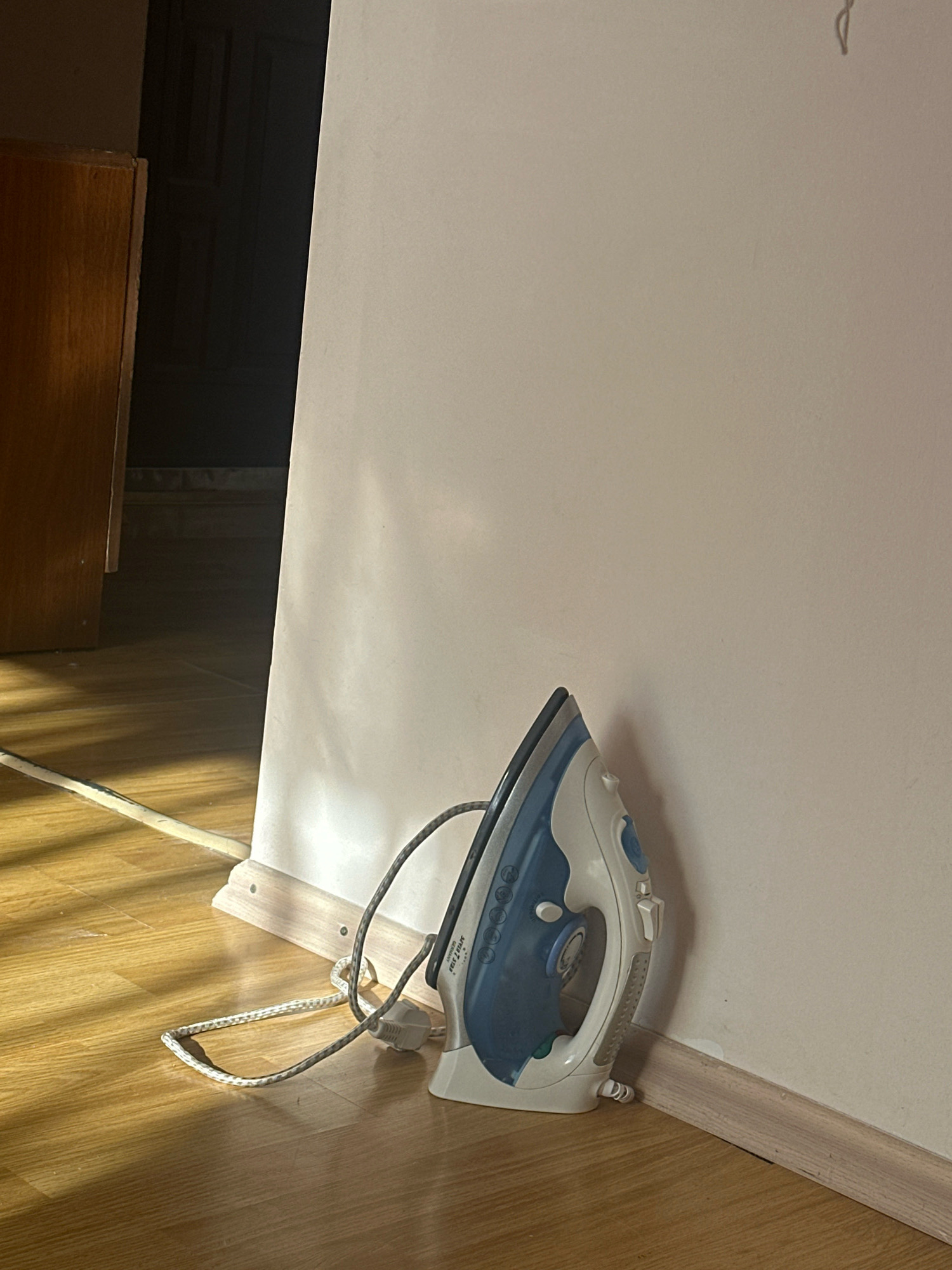 An electric iron, white and blue with a gray cord, rests on a light-colored wooden floor, leaning against a light-colored wall. The cord lies loosely on the floor. Sunlight streams in from the left, casting diagonal shadows across the floor. To the left, a dark doorway and a portion of a wooden cabinet are visible. The scene is indoors, likely in a residential setting in Gyumri, Armenia, given the context of the query, although the image itself does not provide explicit geographic markers. There are no people present, and no actions are depicted beyond the iron being stationary. The environment appears to be a room with wooden flooring and a neutral-colored wall.