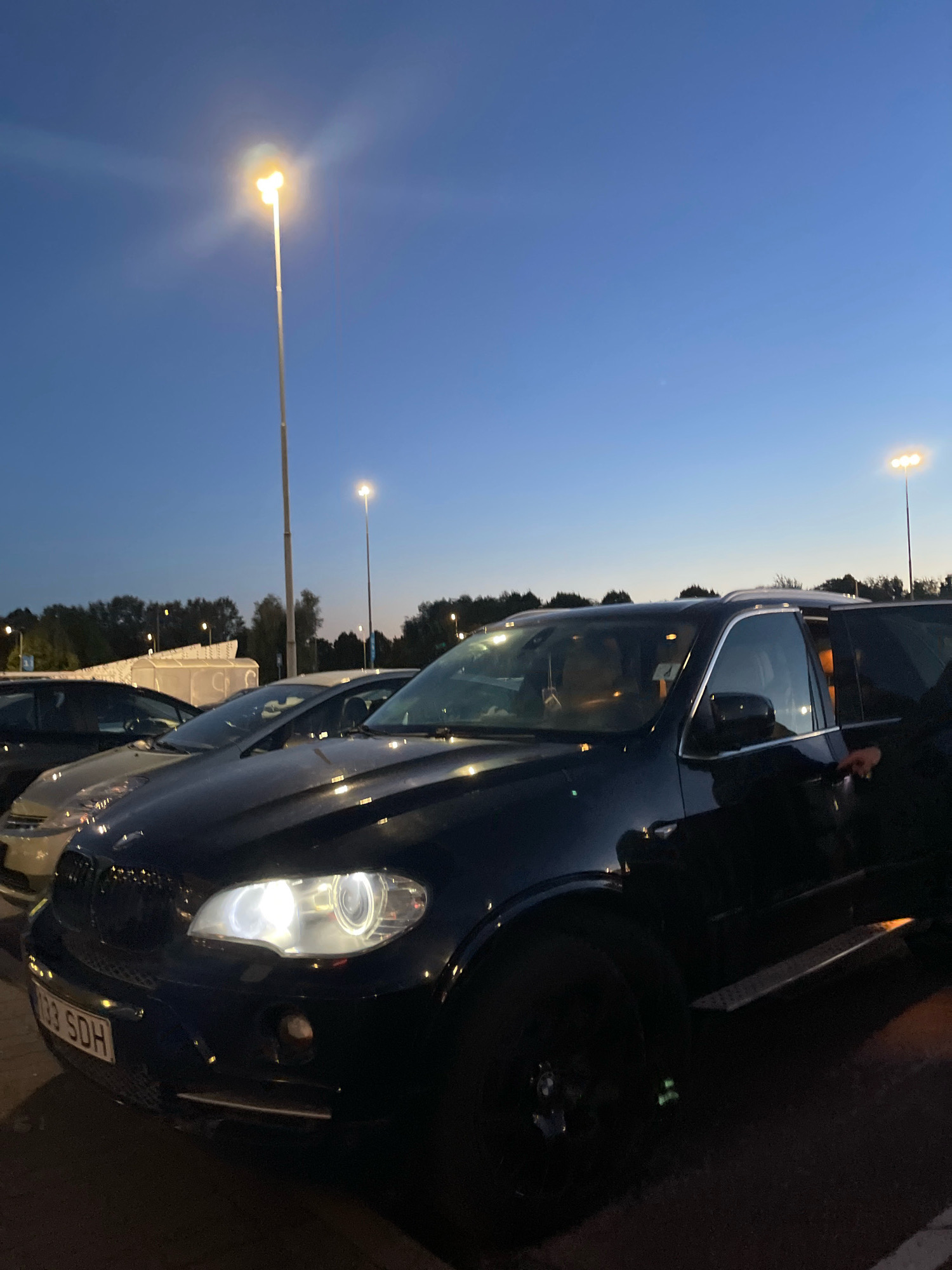 The image shows a dark-colored BMW X5 parked in an outdoor lot with streetlights in the background. The sky is a deep blue, indicating it is dusk or early evening. The car's headlights are on, illuminating the front of the vehicle and the license plate, which reads "133 SDH". A person's hand is visible reaching for the open driver's side door. In the background, other cars are parked, and trees line the horizon. The environment appears to be a parking lot, possibly at a shopping center or public facility, given the presence of multiple streetlights. The image was taken in Tallinn, Estonia.