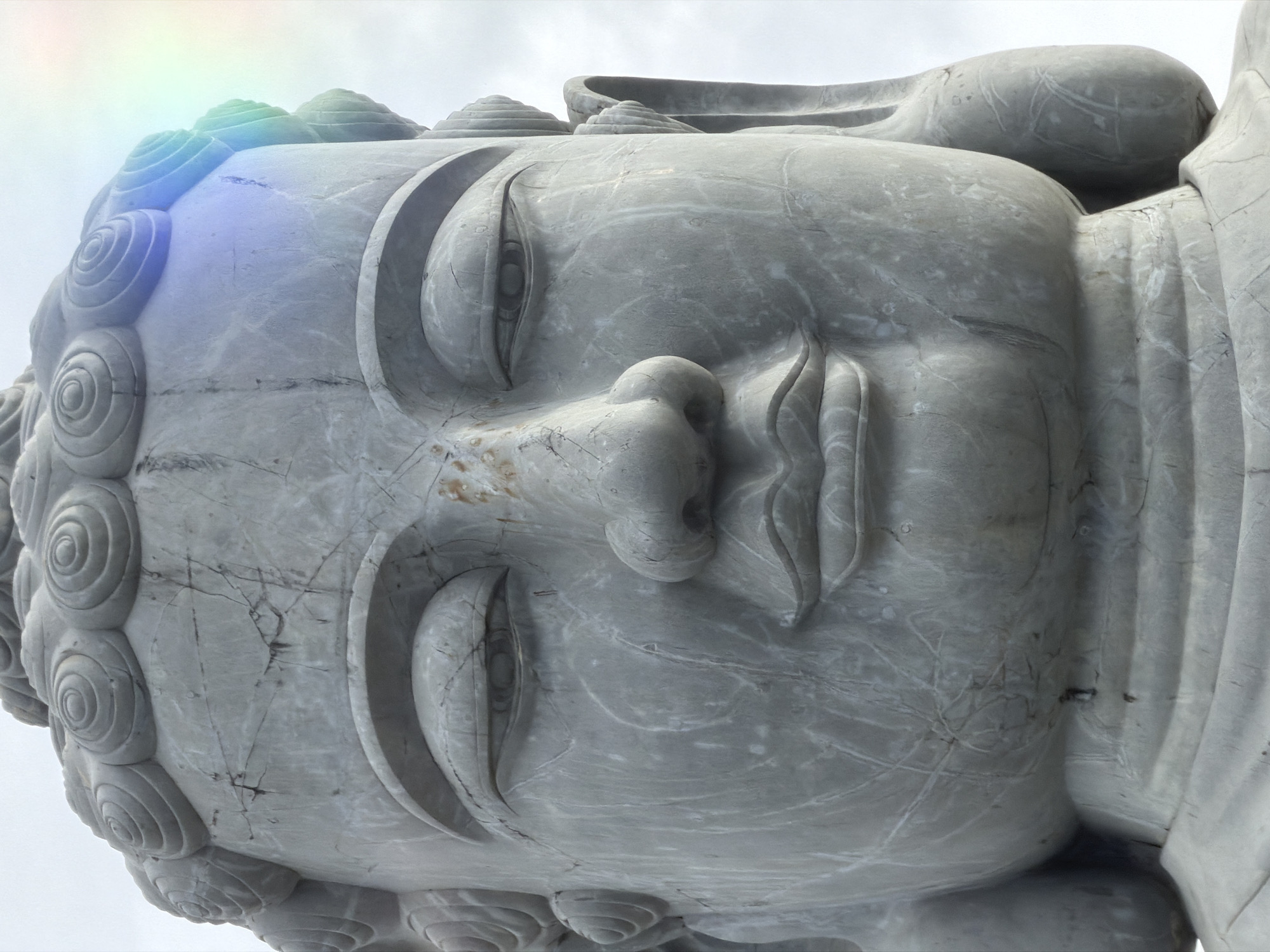 A close-up, low-angle, eye-level shot depicts the face of a gray marble Buddha statue. The statue's features are detailed, showing closed eyes, a small nose, and closed lips. The hair is styled in small, spiraled coils. A faint rainbow-like effect is visible at the upper-left corner of the image. The statue shows signs of weathering and age, with cracks and variations in the stone's color. The sky in the background is overcast. This statue is located in the city of Đà Nẵng, Vietnam.