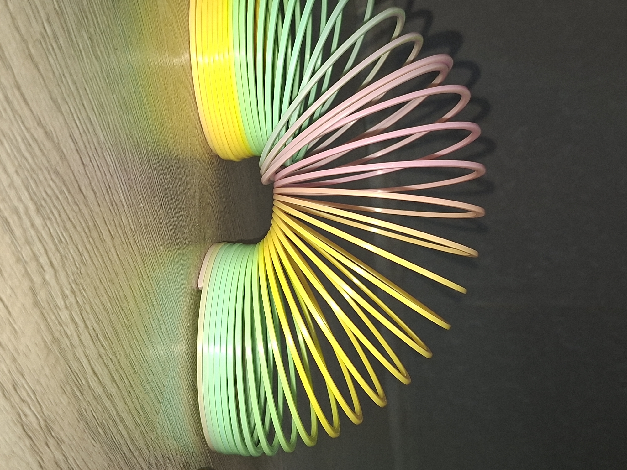 A colorful plastic slinky toy is partially extended on a two-toned surface, half of which appears to be wood grain and the other half a solid dark color. The slinky is a gradient of colors transitioning from green to yellow on one end, extending into a pink hue, and then back to yellow on the other end. The location and the presence of people or actions cannot be determined from the image.