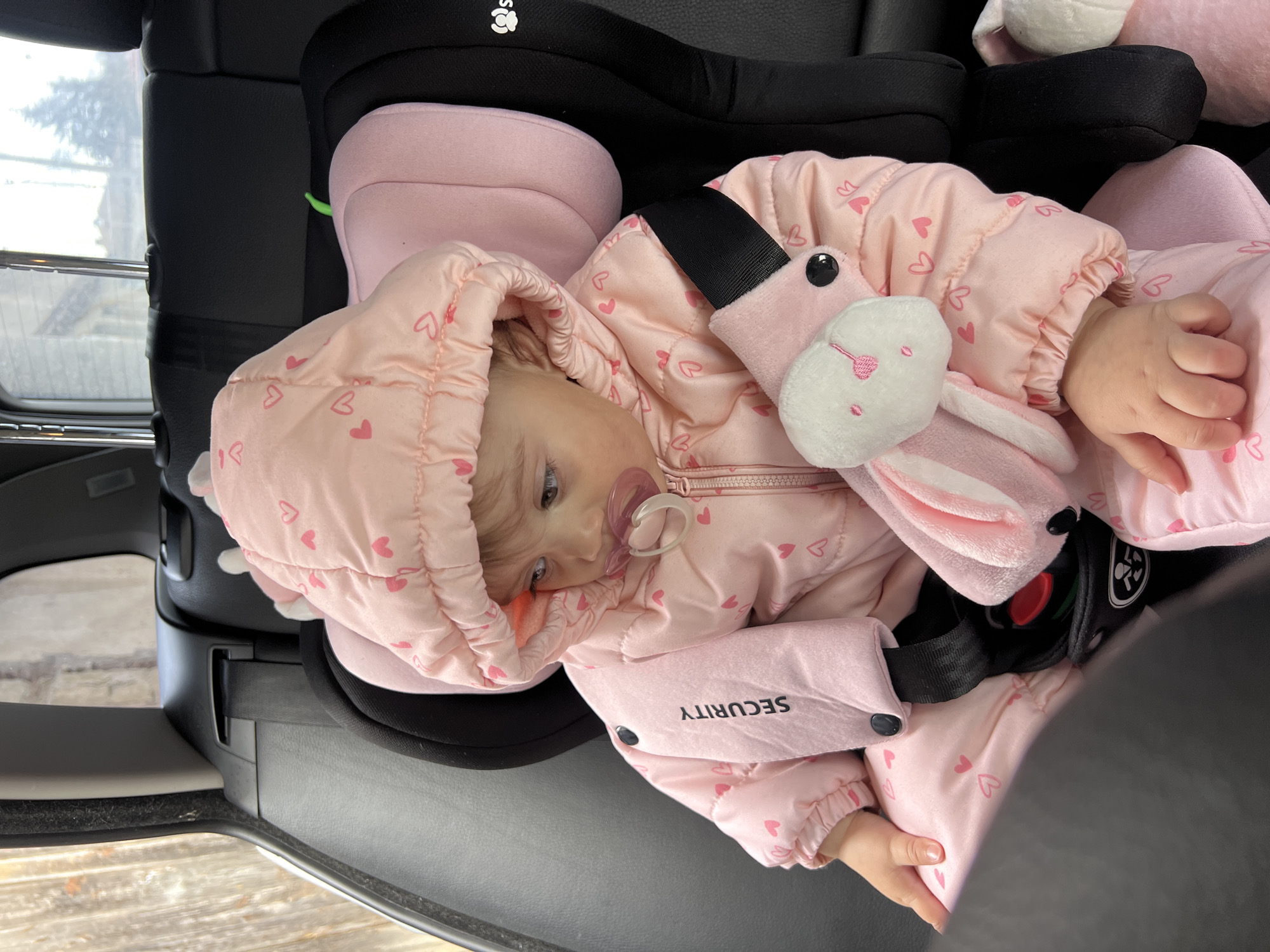 A baby wearing a pink, heart-patterned puffer jacket and a pacifier is secured in a car seat. A stuffed pink bunny is strapped in with the baby. The interior of the car, including the door frame and window, is visible. The location is Elena, Bulgaria.