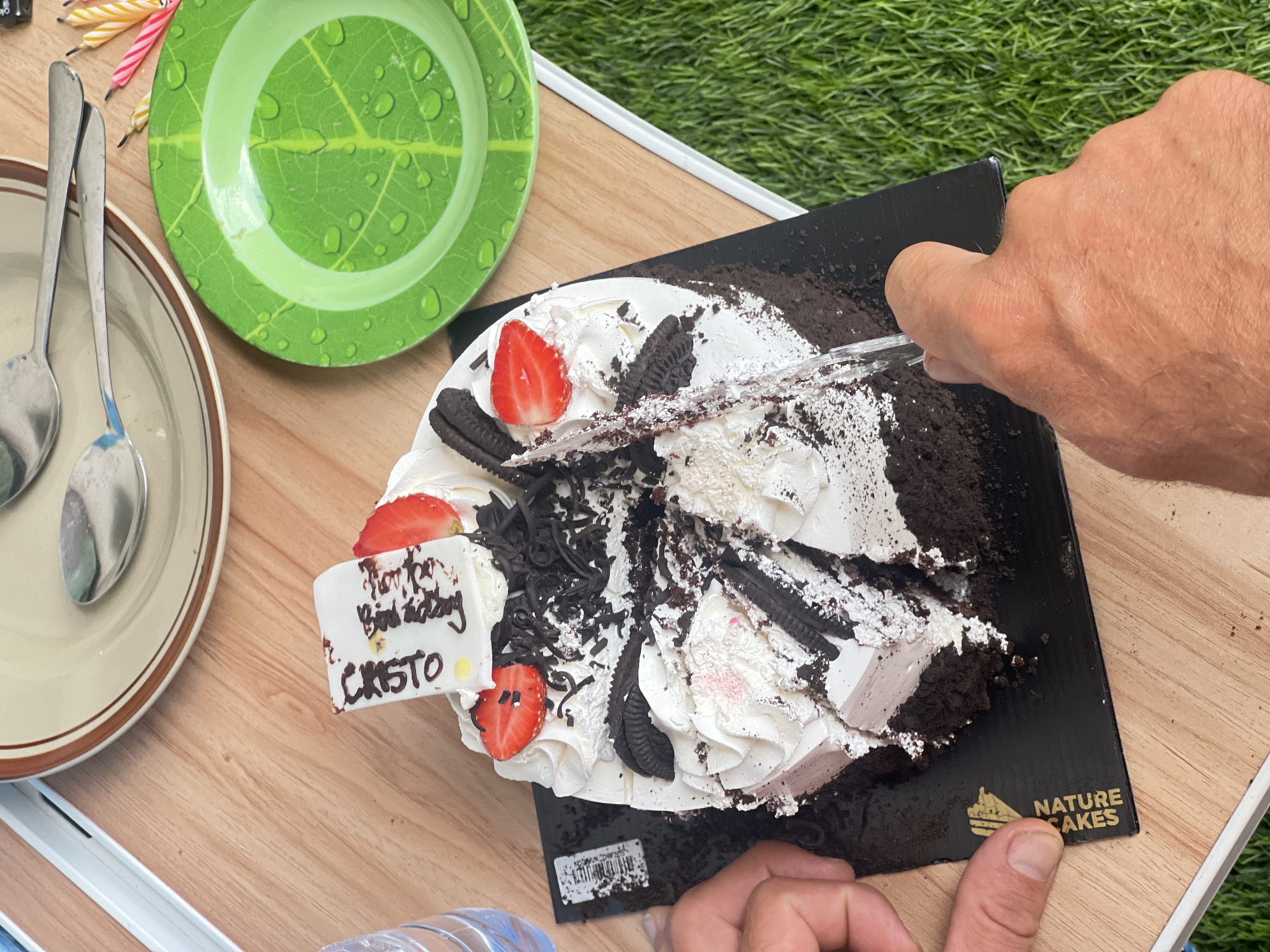 A partially cut chocolate cake with white frosting, strawberries, and Oreo cookies sits on a black cake board on a wooden table outdoors. A knife is mid-cut into the cake. Two plates, spoons, and birthday candles are visible on the table. In the background, a patch of green grass is visible. The scene is likely a backyard or patio in Denpasar, Indonesia, indicated by the presence of outdoor elements and typical Indonesian environmental features. The overall impression is a casual, celebratory gathering.
