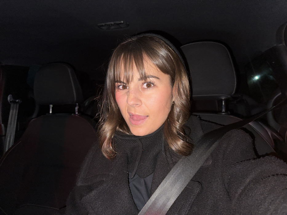 A young woman with brown hair and bangs sits in a car, sticking her tongue out. She wears a black coat over a turtleneck and a black beanie. The car interior is dark, with black seats and a seatbelt across her chest. The photo is taken at night, with only dim lighting. She appears to be alone in the vehicle.