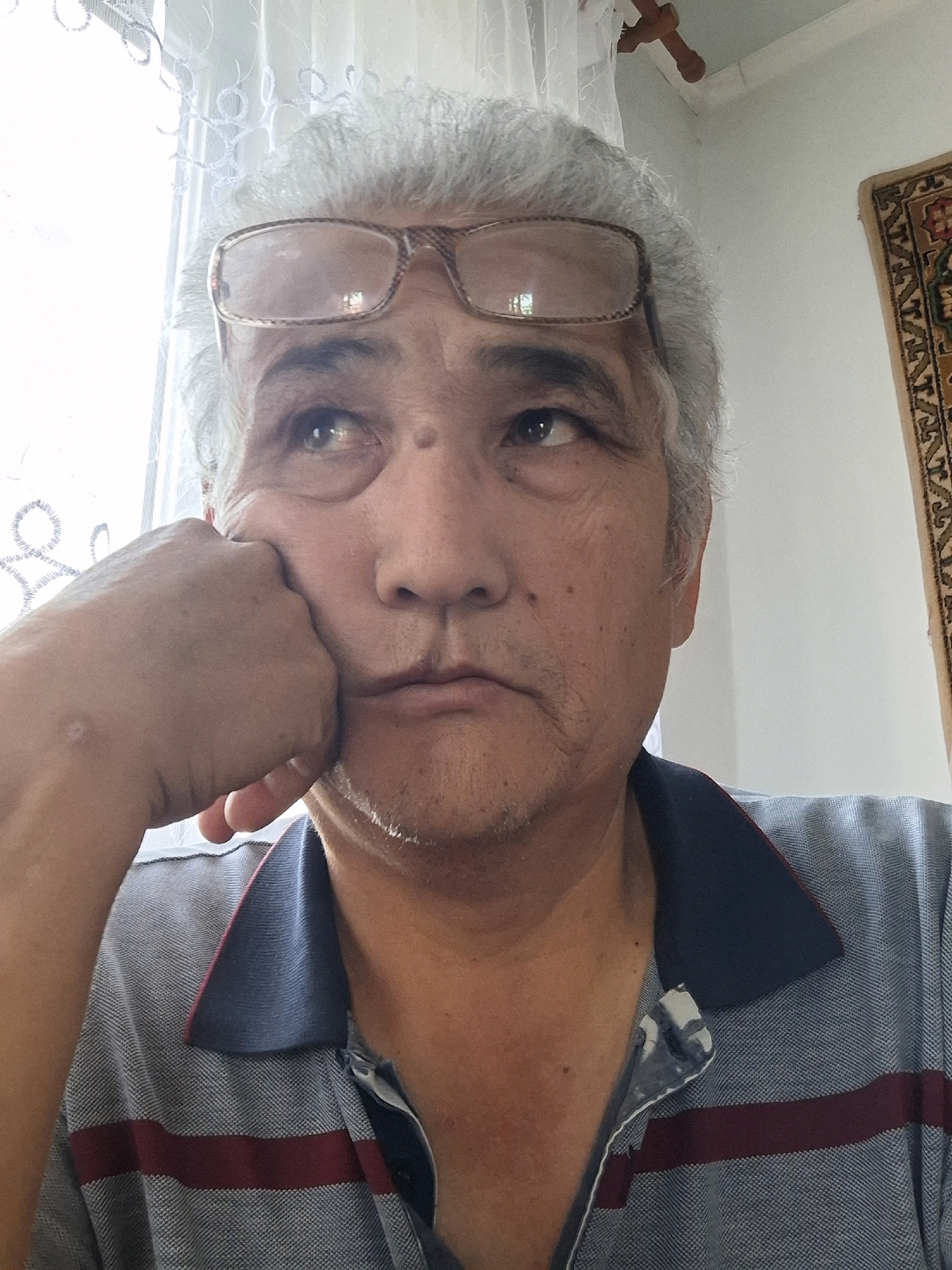 A close-up, eye-level, selfie-style photo shows a middle-aged man with graying hair, wearing glasses and a gray polo shirt with a dark blue collar and maroon stripes, resting his chin on his fist. He appears to be looking upwards and to his left, with a pensive or bored expression. The background is a dimly lit room with a sheer curtain to the left, a white wall, and a small portion of a decorative rug hanging on the wall to the right. The lighting suggests an indoor setting, possibly a home, with natural light coming from the left. The overall impression is that of a candid moment captured in a domestic environment. No specific identifying landmarks of Almaty, Kazakhstan are visible.