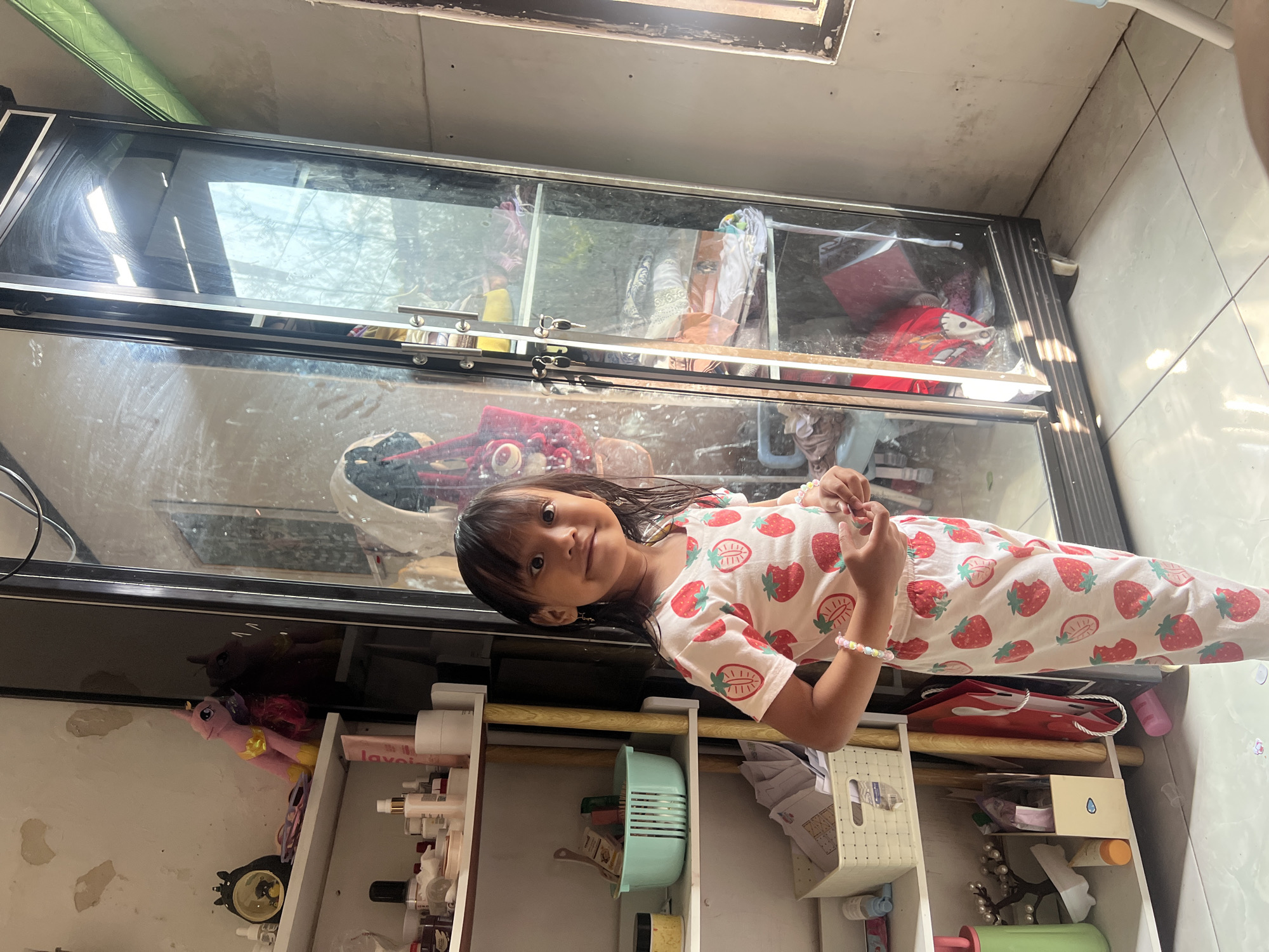A young girl wearing strawberry-patterned pajamas stands in a room in Kecamatan Pati, Indonesia. She is positioned to the left of a large, reflective cabinet with glass doors. The cabinet contains shelves filled with various items, including clothing and bags. To the girl's left is a multi-tiered shelf unit holding toiletries, a light blue basket, and other miscellaneous objects. Sunlight streams into the room, casting shadows on the floor and the cabinet. The walls appear slightly aged and textured. The girl is looking towards the camera with a slight smile.