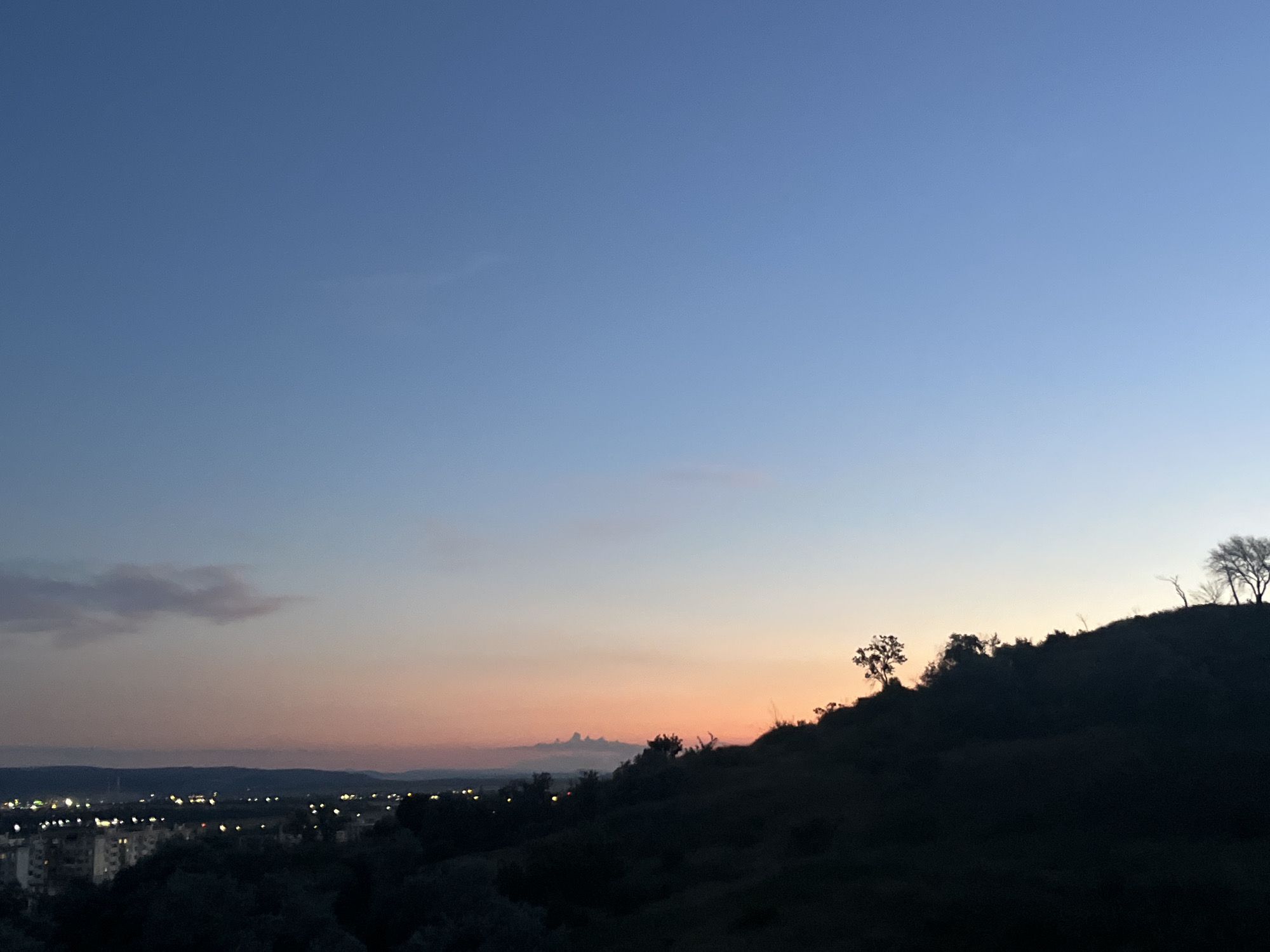 The image captures a twilight scene over the city of Varna, Bulgaria, likely taken from an elevated viewpoint. The sky transitions from a deep blue at the top to lighter shades of blue and then to soft hues of pink and orange near the horizon, indicating sunset or sunrise. A faint cloud formation is visible in the upper left quadrant of the sky.
In the mid-ground, a silhouetted hill dominates the right side of the frame, dotted with several small, bare trees. The city lights of Varna are visible in the distance on the left, twinkling amongst buildings. A dark, wooded area or foliage is present in the foreground, partially obscuring the lower portion of the cityscape. The overall impression is one of peaceful dusk settling over the urban landscape.