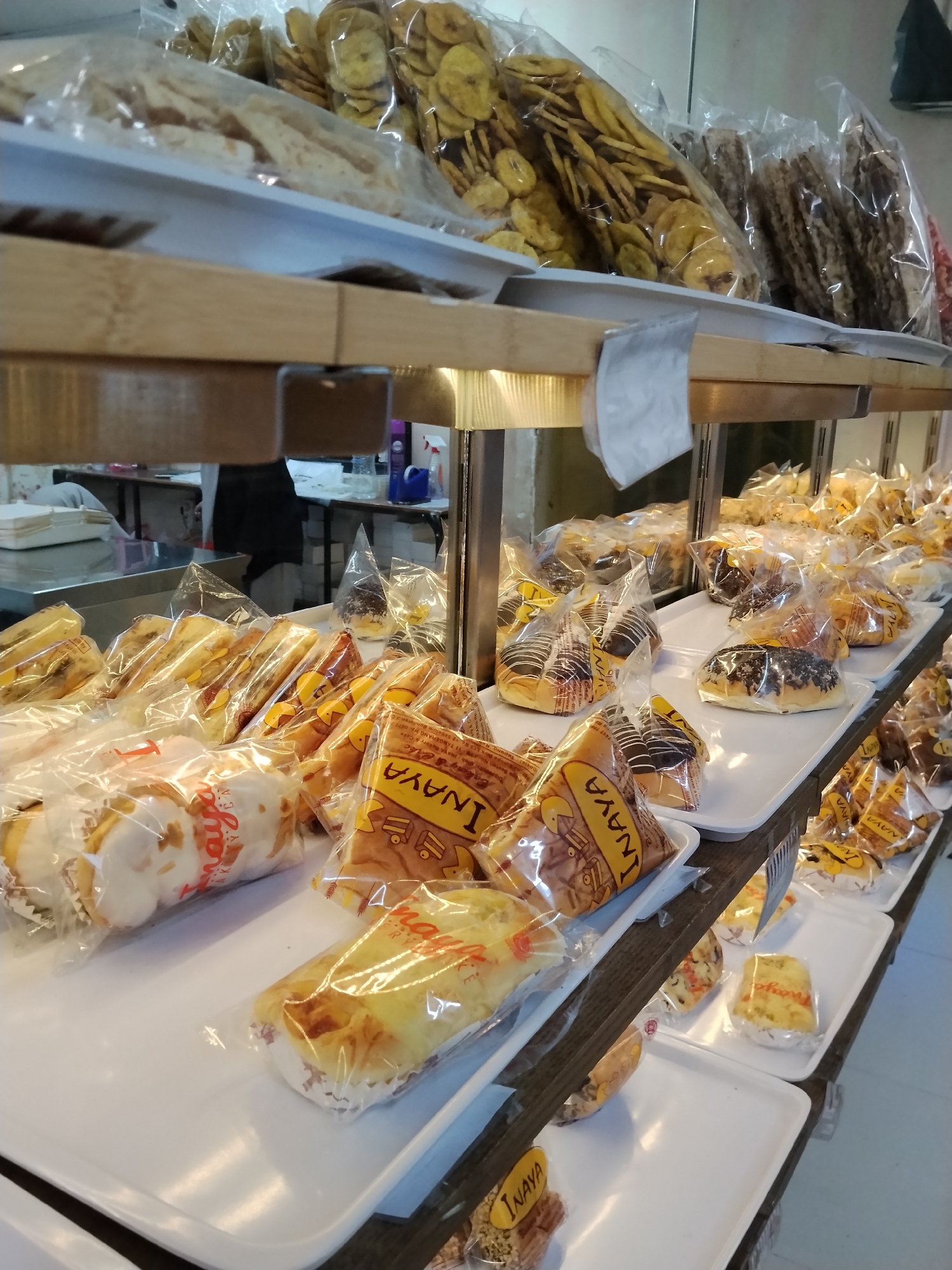 This is a close-up, eye-level, medium shot of a bakery display in Ciruas, Indonesia. The focus is on multiple shelves stacked with various types of bread and snacks, all individually packaged in clear plastic. The lighting suggests an indoor environment, possibly a brightly lit storefront, with a wooden shelf structure supporting the display.
On the top shelf, there are bagged items that appear to be chips or crackers, some with a reddish hue and others with a lighter, more golden color. The middle shelf features an assortment of baked goods, including round pastries with chocolate frosting, elongated bread rolls, and square-shaped cakes or brownies. Many of these are also in clear plastic packaging, some with visible branding or text in yellow and red. The bottom shelf is partially visible, displaying more packaged pastries and some items that resemble small loaves of bread.
The overall impression is one of abundance and variety within a typical Indonesian bakery setting. There are no visible people in the immediate foreground or on the displayed shelves. The background is blurred, but hints of a counter and possibly other display cases can be discerned. The scene is clean and organized, with white plastic trays holding the packaged items, creating a neat presentation.
