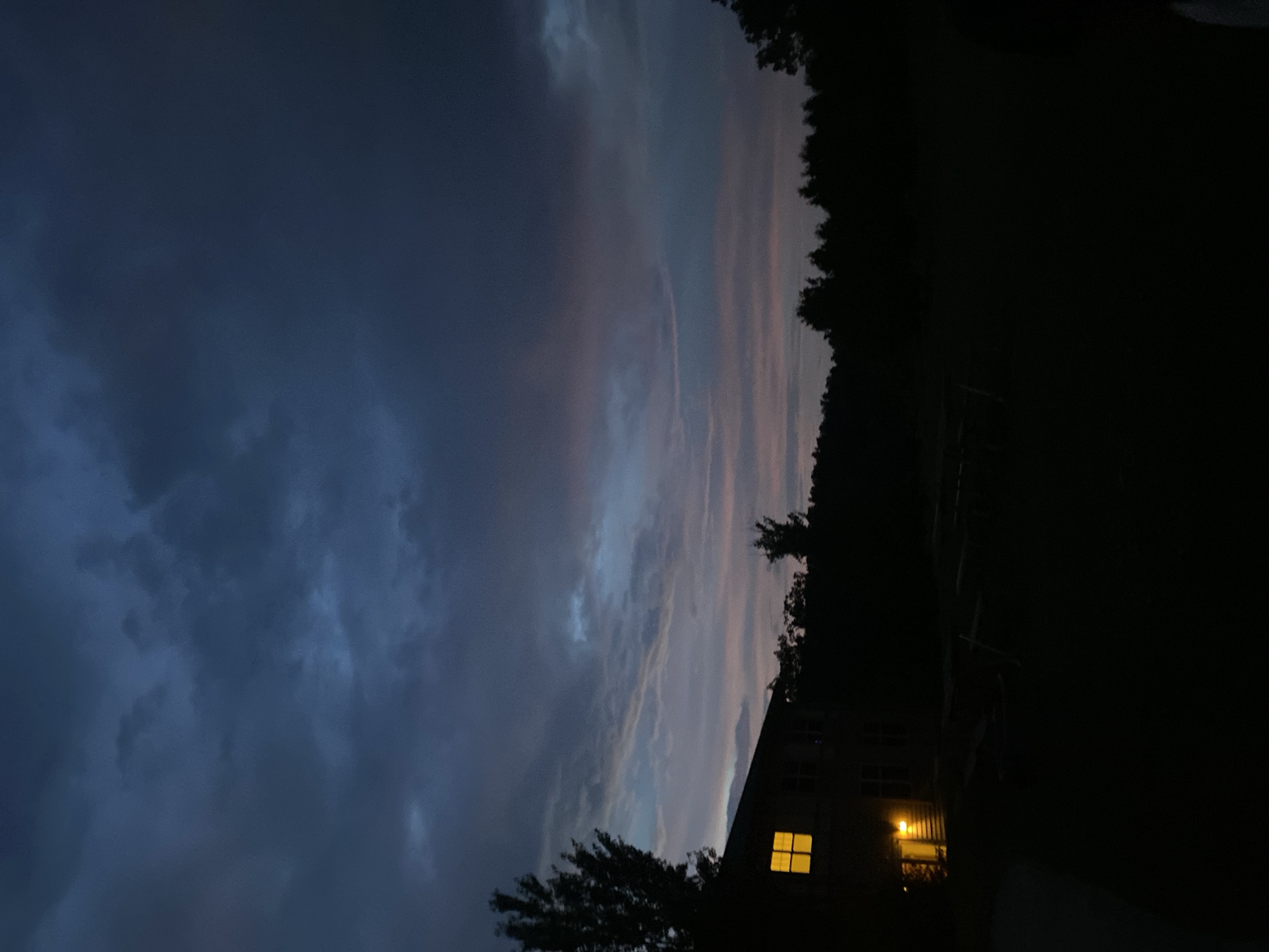 In Washington Township, United States of America, this image captures a dimly lit scene at dusk or night. A two-story house, partially visible on the left, has several windows illuminated with a warm, yellow light. A large tree stands to the left of the house, silhouetted against the sky. The foreground consists of a dark, grassy yard with indistinct shapes that could be outdoor furniture or equipment. A dense line of trees forms a dark backdrop across the midground. The sky is a deep, moody blue, with heavy clouds that are tinged with subtle shades of pink and purple, suggesting the last vestiges of daylight. The overall atmosphere is one of quietude and the transition from day to night.