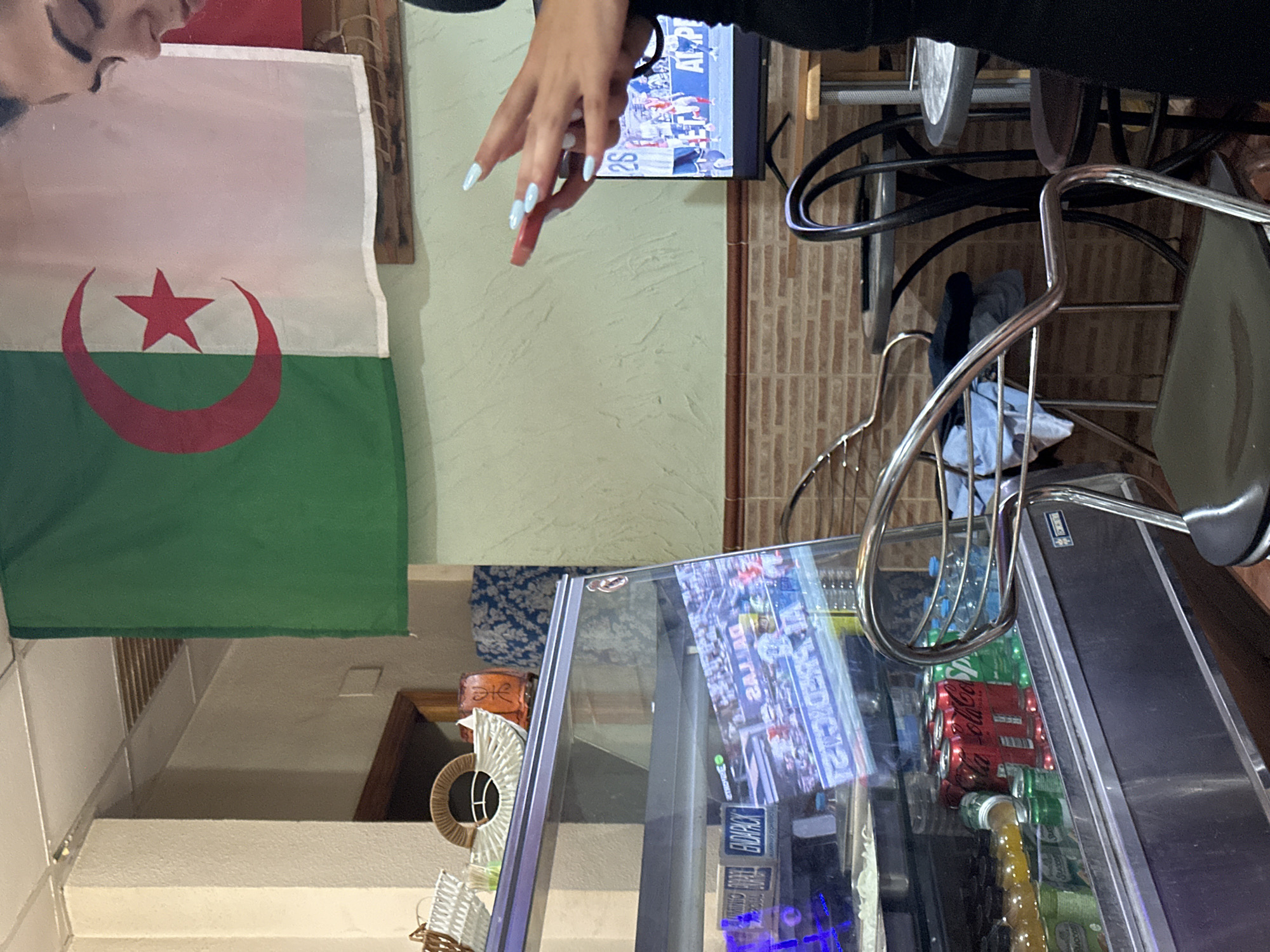 The image depicts the interior of a casual establishment, possibly a cafe or small restaurant, located in the city of Alicante/Alacant, Spain. A national flag, likely that of Algeria, hangs prominently on a wall. In the foreground, a glass display case is filled with drinks, including Coca-Cola and Sprite, and appears to contain some packaged items. A young woman, partially visible from the side, is holding a red object, possibly a phone or remote, and looking downwards. Behind her, a television screen displays a sports event. The environment features typical cafe furnishings, such as tables and chairs, and tiled walls. The overall impression is of a relaxed, everyday setting.