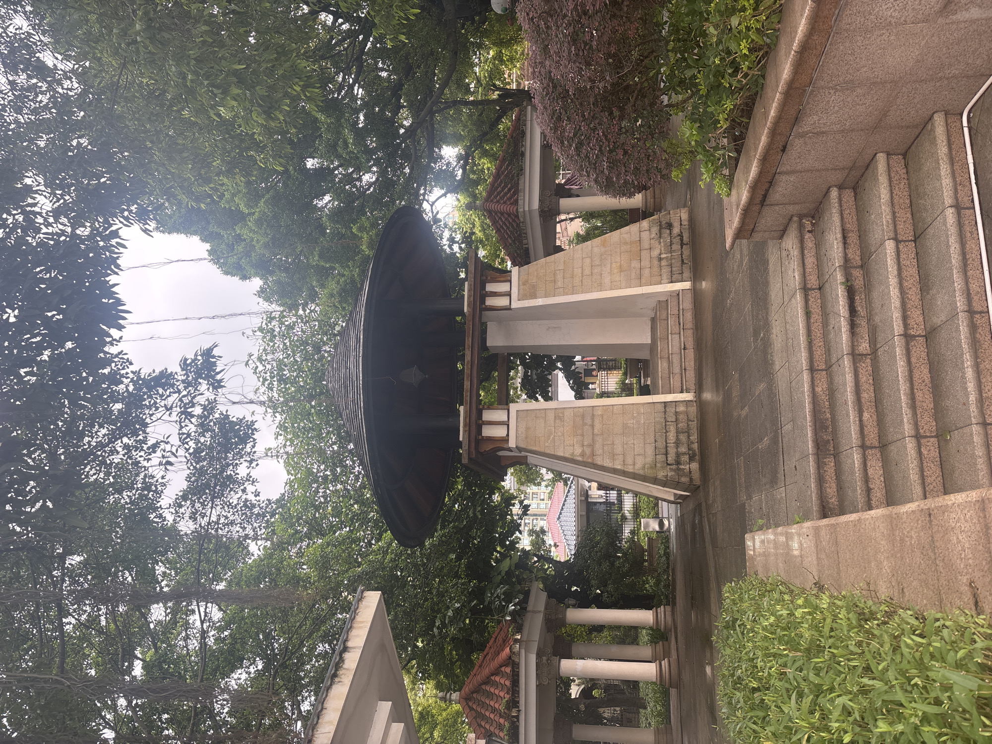 This is a photograph of a gazebo in a park in Nanhai District, China. The gazebo is octagonal with a dark, tiled roof and a stone base. It is surrounded by trees and lush green foliage. In the foreground, there are stone steps leading up to the gazebo. To the left of the gazebo, there is another smaller structure with a red tiled roof and white columns. The ground is paved with stone tiles and appears to be wet, suggesting recent rain. The sky is overcast. There are no people visible in the image.