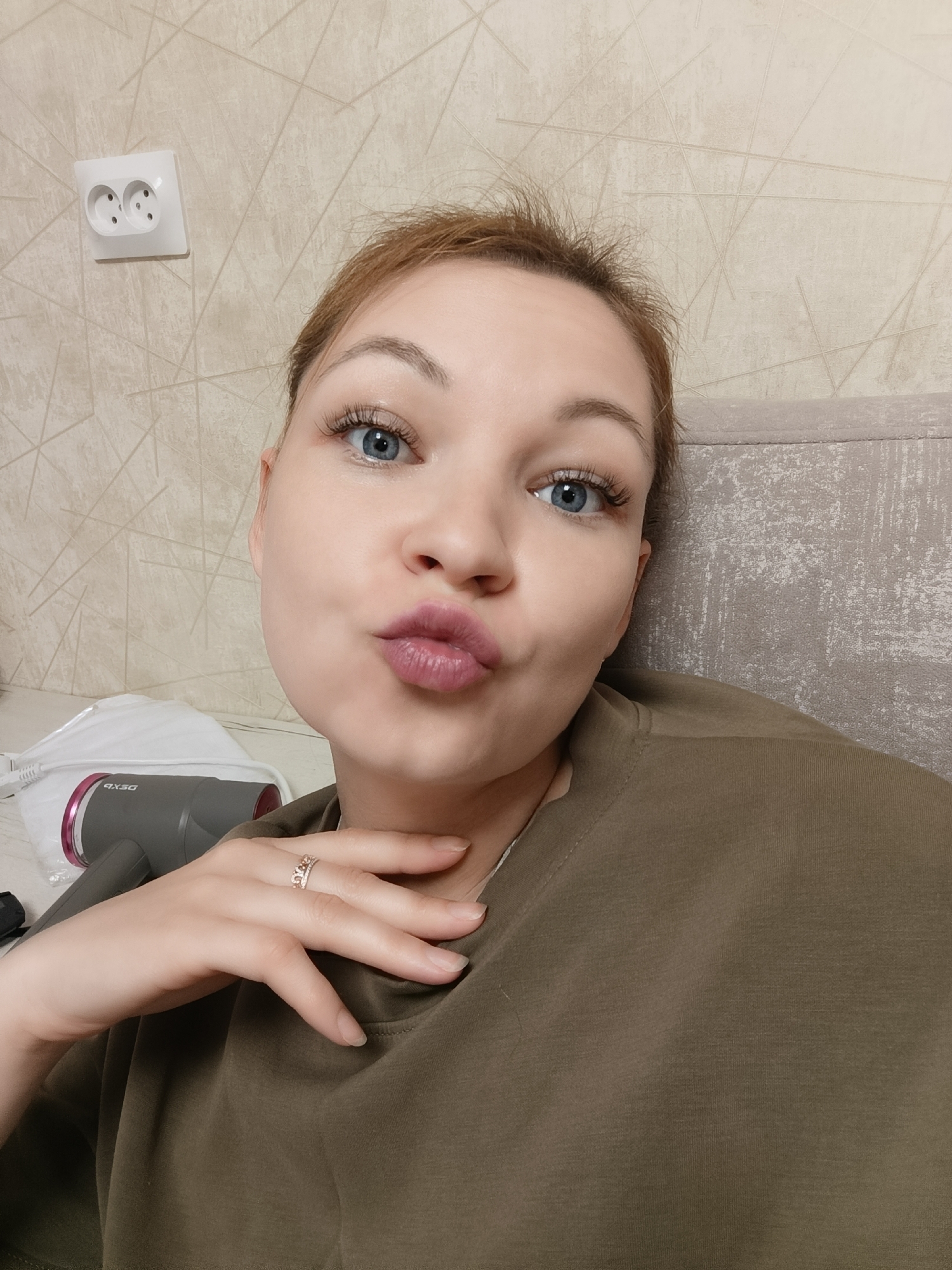 A woman is taking a selfie in an indoor setting. She has blonde hair pulled back, blue eyes, and is puckering her lips. She is wearing a brown t-shirt and a ring on her finger. In the background, there is a wall with a patterned wallpaper and an electrical outlet. There is also a gray hair dryer and a white mask on a surface to her left. The setting appears to be a bedroom or living area. The image does not contain information that allows for localization in Chaikovsky, Russia.