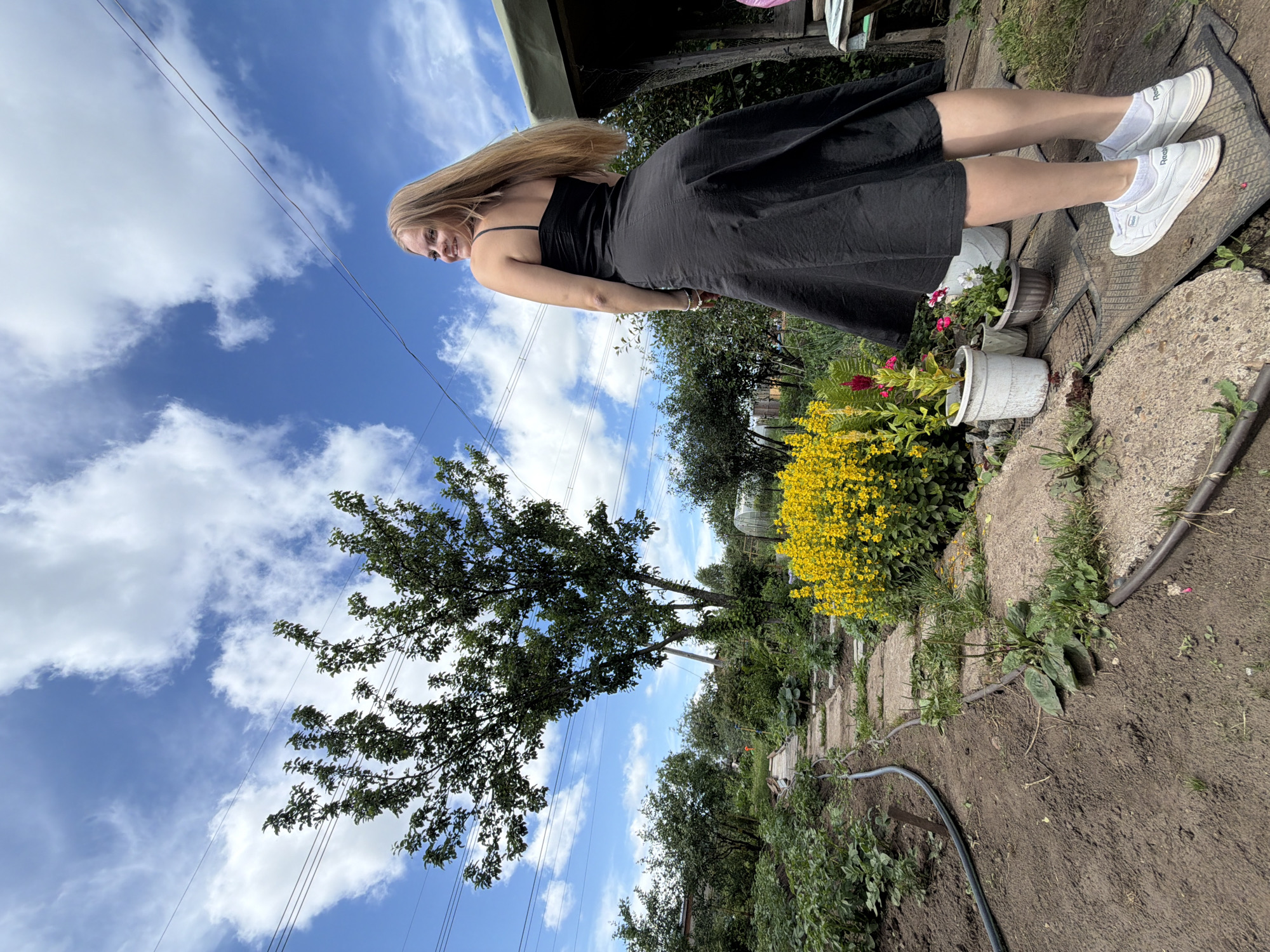 In Chaikovsky, Russia, a young woman with long blonde hair, wearing a black sundress and white sneakers, stands in a garden on a sunny day. She is looking over her left shoulder and smiling at the camera. The garden is filled with lush green plants, a path made of concrete slabs, and a cluster of bright yellow flowers. In the background, a large tree with green leaves stands tall against a bright blue sky with scattered white clouds. Power lines are visible crisscrossing the sky. To the right, a covered structure, possibly a shed or greenhouse, is partially visible.