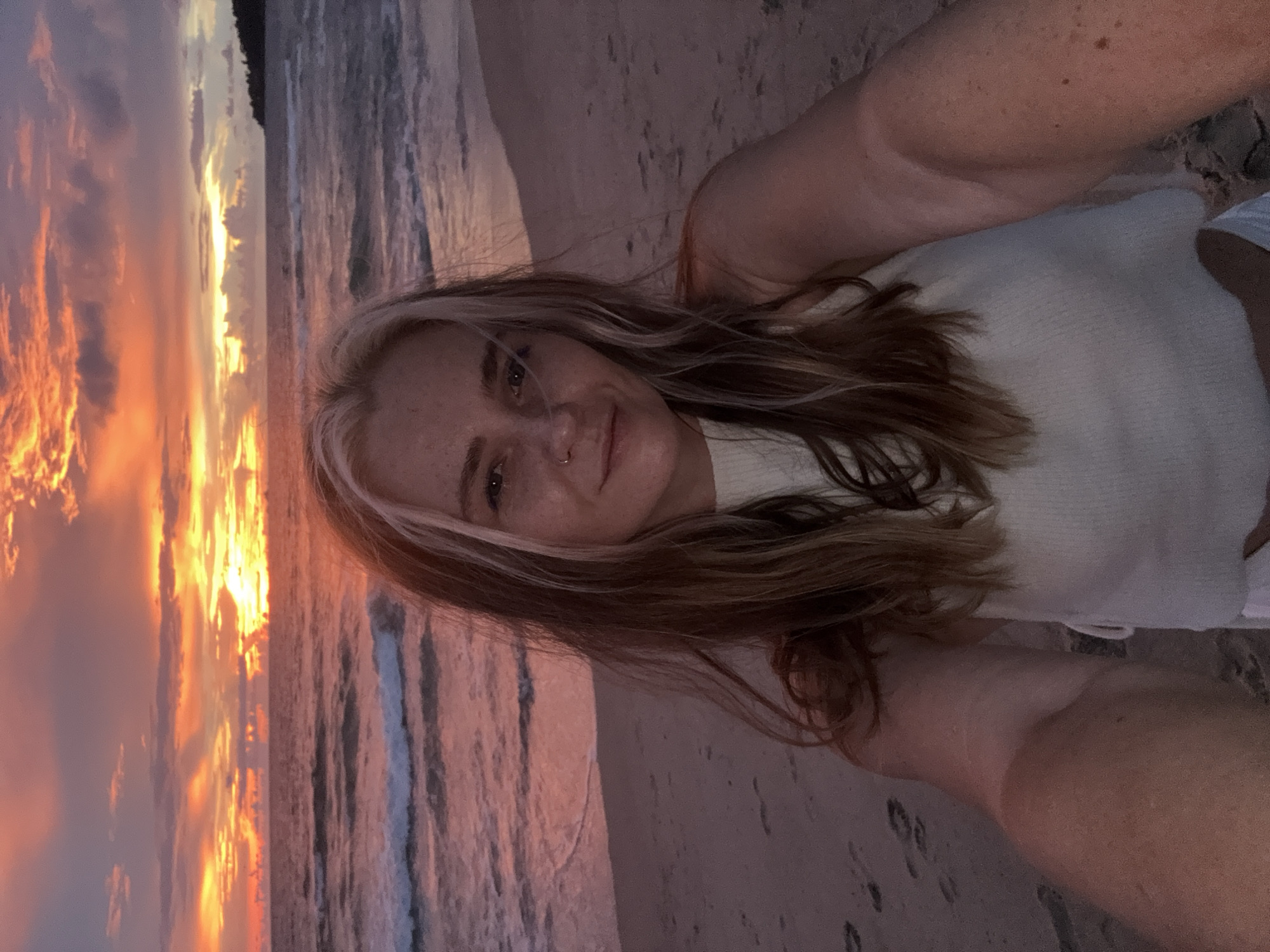 A woman with blonde streaks in her brown hair takes a selfie on a beach at sunset. The sky is ablaze with orange and pink hues, with dramatic clouds. The ocean is calm with gentle waves lapping the shore. The sand is light brown with footprints visible. The woman is wearing a white top and her left arm is extended, holding the camera. She is smiling at the camera. In the background, a distant headland is visible on the right side of the frame. The overall impression is serene and picturesque. The location is likely a beach in Choeng Thale, Thailand, given the typical sunset colors and coastal scenery.