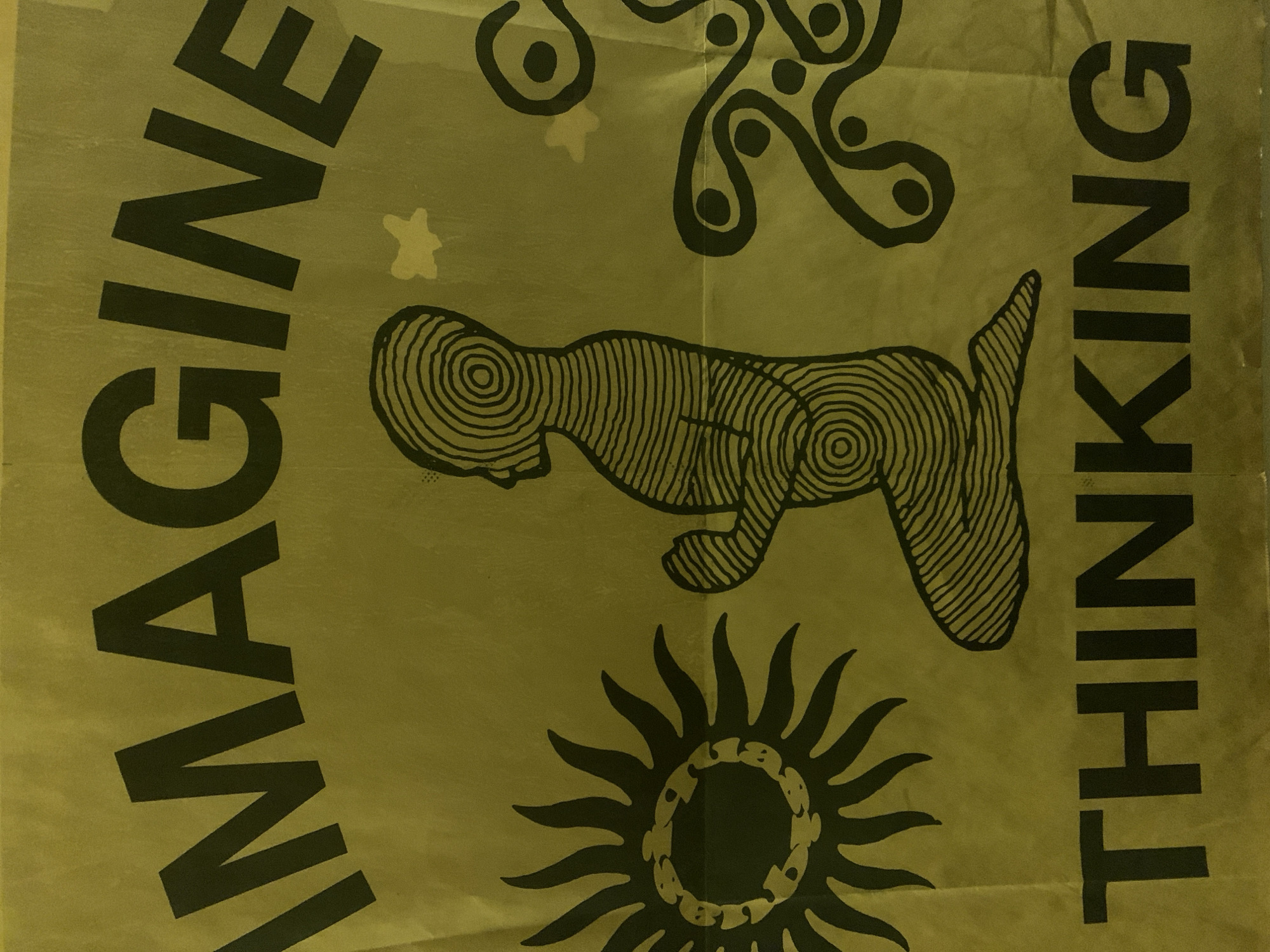The image shows a poster or print in a yellowish-green hue. The words "IMAGINE" are written vertically on the left, and "THINKING" on the right, both in black capital letters. In the center of the poster, there are various black designs. These designs include a quadrupedal figure with concentric circles forming its body, a sun with radiating rays, and abstract swirling shapes. There are also a couple of asterisk-like shapes scattered in the background. Without additional information, it is impossible to pinpoint the exact location within Barcelona, Spain.