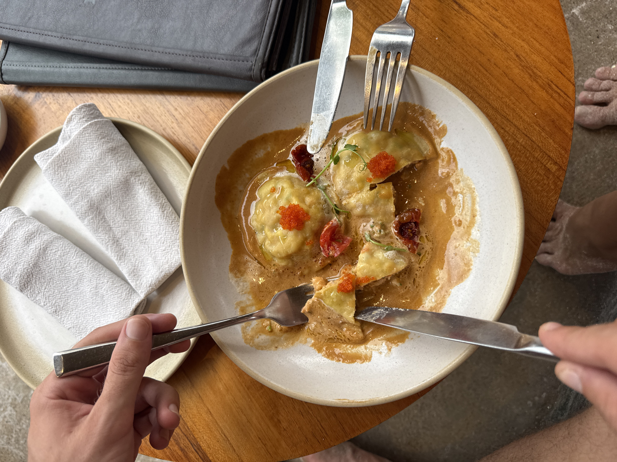 An overhead shot captures a person eating a meal, likely at a cafe or restaurant, with their bare feet visible on the ground. The central focus is a white, shallow bowl containing several pieces of ravioli or similar filled pasta in a creamy, reddish-brown sauce. The food is garnished with bright orange roe, small pieces of dried red tomatoes, and tiny green microgreens. A fork and knife are positioned on the food, and another fork and knife are held by the person's hands, actively cutting into a piece of the pasta.
To the left of the main dish, a smaller beige plate holds two neatly folded white napkins with a subtle woven pattern. In the upper left corner, a dark grey, possibly leather, wallet or folder rests on a wooden table surface. The table surface is a warm, medium-brown wood.
The person's hands, with clear fingernails, are visible in the foreground, holding the cutlery. To the right, at the bottom of the frame, parts of the person's bare legs and feet are visible on a lighter grey, textured floor surface, which appears to have some light dust or sand on it. The lighting suggests it is daytime. No text is visible in the image.