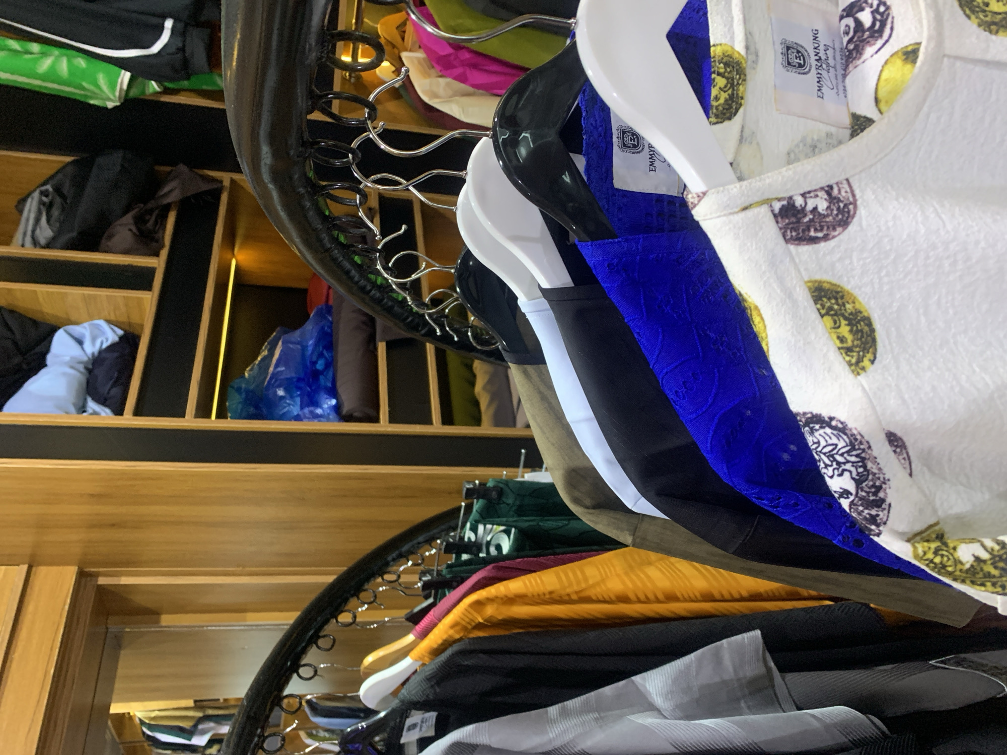 A retail clothing display features several garments hanging on racks and shelves in what appears to be a store. The clothing includes items in various colors such as black, yellow, white, and blue, with some garments exhibiting patterned designs. In the background, shelves are visible, some containing folded clothes and plastic-wrapped items. The overall environment suggests a commercial setting for apparel sales. The location is identified as Gura Top, Nigeria.