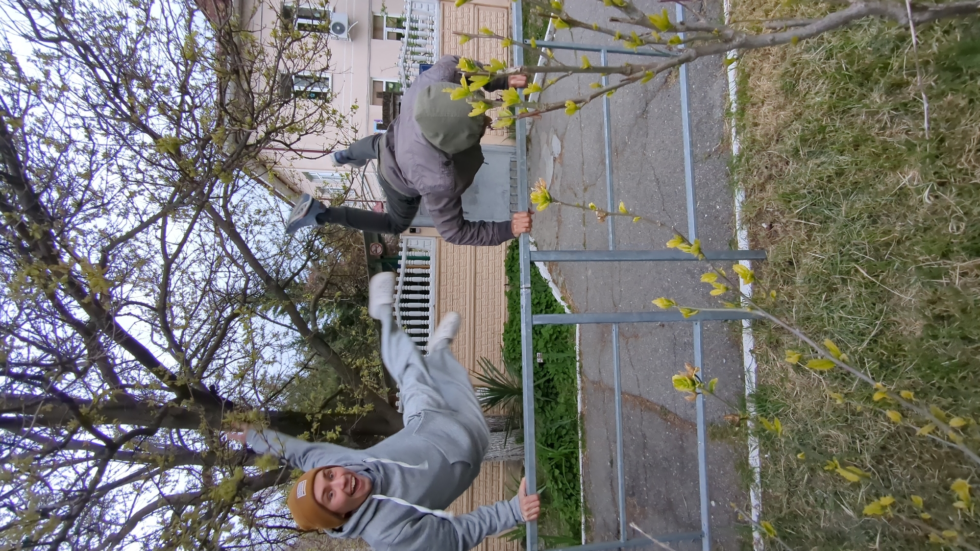 Two young men are engaged in an acrobatic or parkour activity on a metal fence in an outdoor setting, likely in Gelendzhik, Russia. The image is taken from a low angle, looking upwards through tree branches.
One man, wearing a grey hooded tracksuit and a brown beanie, is in an inverted position, hanging with his legs extended upwards and his head down. He is holding onto the bottom bar of the metal fence with one hand and has his other hand raised, giving a thumbs-up gesture. He has a wide smile on his face, indicating enjoyment or excitement. His white sneakers are visible.
The second man, wearing a grey hooded jacket and dark pants, is positioned above the fence, appearing to be vaulting over it or balancing on its top bar. His legs are bent, and his hands are gripping the fence. His face is not clearly visible.
The scene takes place during the daytime, under an overcast sky which appears bright white/light grey. The trees in the foreground and background are in early spring, with bare branches beginning to sprout small, light green leaves. Behind the fence and the men, there is a paved area, likely asphalt, and a strip of grass. In the background, a light-colored building with balconies, windows, and an air conditioning unit is visible, suggesting a residential or hotel environment. The metal fence is a simple, rectangular structure. No visible text is present in the image.