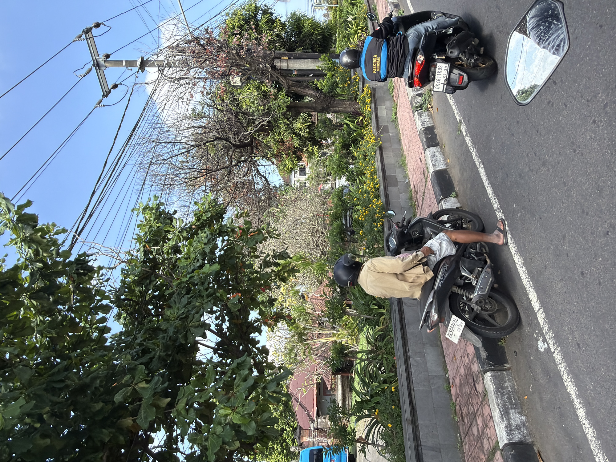 In Blahbatuh, Indonesia, two individuals are riding motorcycles on a road on a sunny day. The first motorcyclist, seen from the rear, is wearing a light-colored jacket and shorts, with a black helmet. The motorcycle is dark-colored and has a license plate reading "DK5068KAL". The second motorcyclist, also seen from the rear, is wearing a blue and black jacket with "PASKIBRA SDENSAGI" printed on it, and jeans, also with a black helmet. Their motorcycle is dark-colored and displays a license plate reading "DK4611KBS" and "07-29". The road is paved with a white line marking the lane. To the left of the road is a sidewalk made of red bricks and a gray curb. Beyond the sidewalk is a landscaped area with lush green trees, some with bare branches, and bright yellow flowers. In the background, traditional Indonesian buildings with red roofs are visible amongst the foliage, along with a blue vehicle. Overhead, utility poles with numerous wires crisscross the bright blue sky.