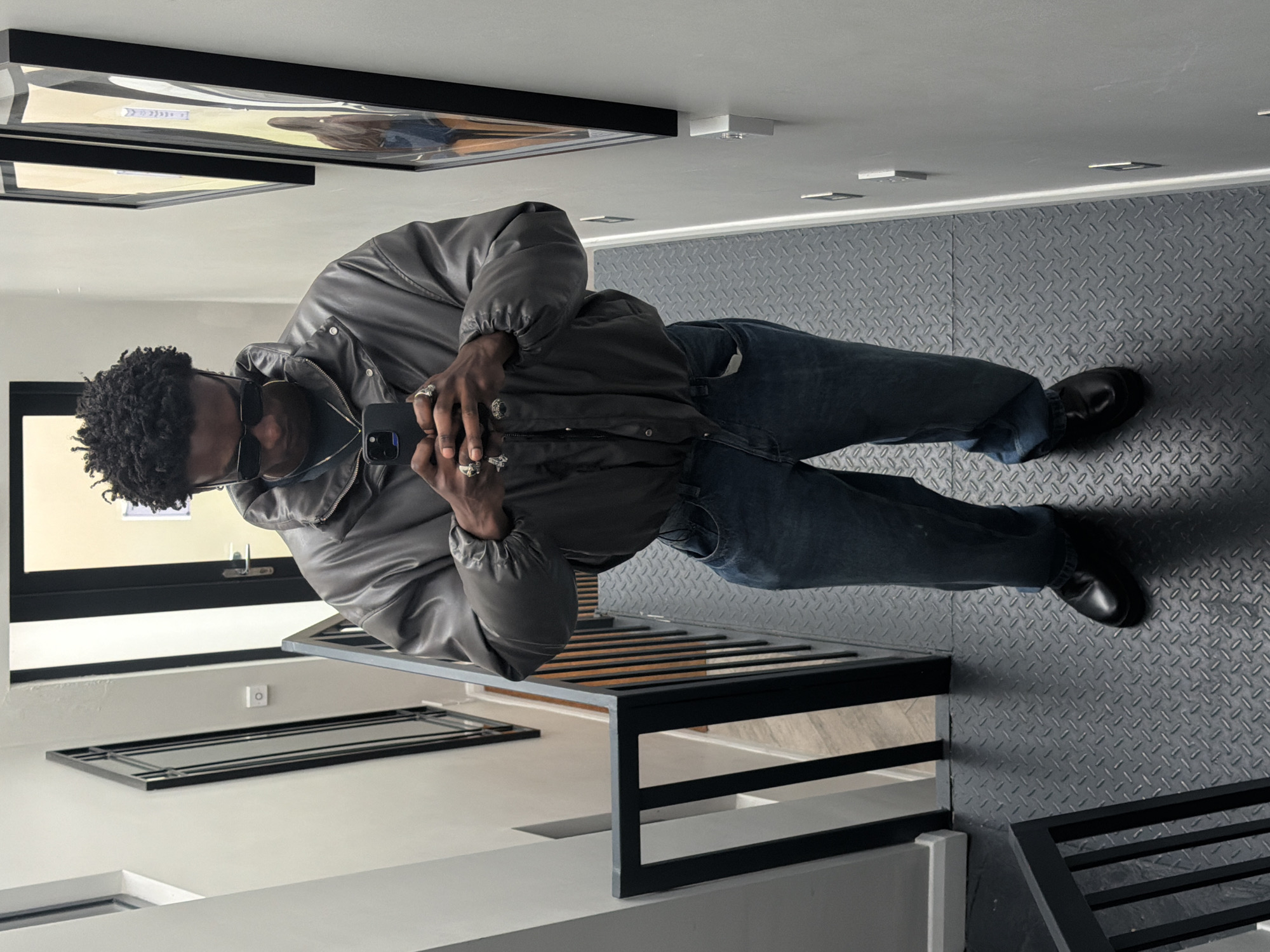 A man is taking a selfie in what appears to be an art gallery or modern building. He is wearing a grey puffer jacket, dark jeans, and black leather shoes. He has dark curly hair and is wearing sunglasses and a face mask. Multiple rings adorn his fingers. He is holding a black smartphone in his hand. The floor is covered in grey diamond-patterned tiling. The wall adjacent to him has a black-framed window and what may be artwork, with a black railing in the foreground. Additional framed artwork appears on the ceiling. The location cannot be determined specifically to London, United Kingdom based on the provided image.