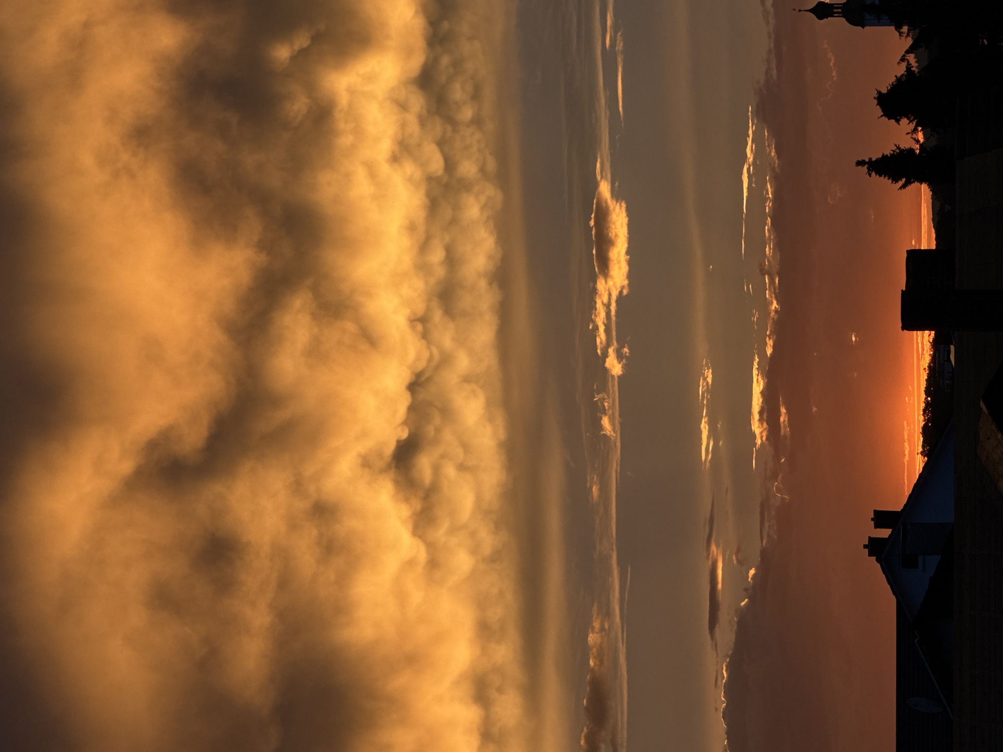The image shows a dramatic sunset over the city of Wettstetten, Germany. The sky is filled with vibrant orange and golden clouds, with layers of darker clouds in between. The sun is low on the horizon, casting a warm glow over the scene. In the foreground, silhouetted against the sky, are the rooftops of houses and buildings, along with several trees and the spire of a church. The overall impression is one of a beautiful and powerful natural phenomenon.