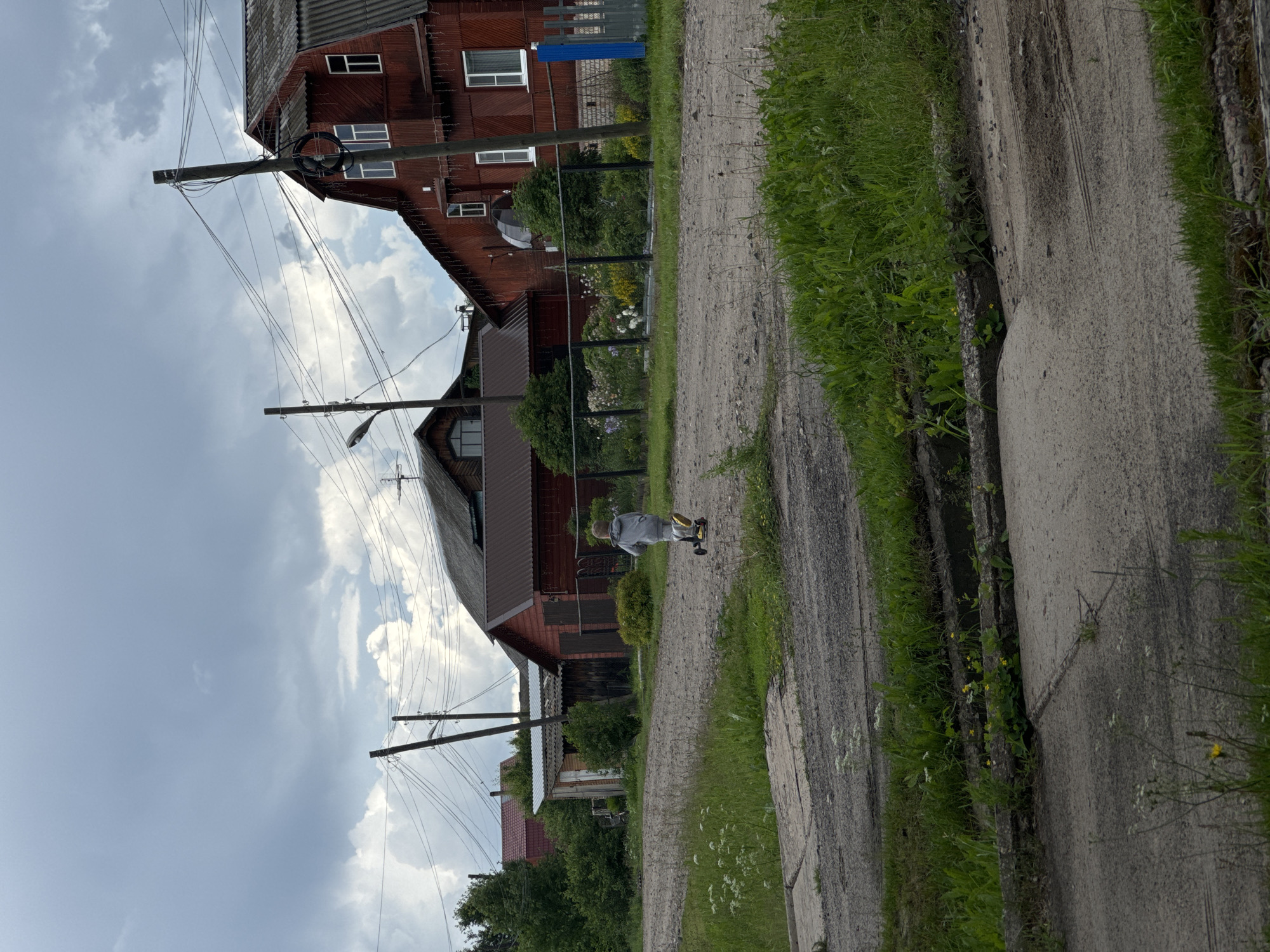 The image depicts a rural street scene in the village of Ponaryevo, Russia, under an overcast sky. A young child, dressed in a grey hooded outfit and yellow shoes, is riding a scooter on a gravel road. Behind the child, a dark red, wooden house with a corrugated metal roof dominates the right side of the frame, featuring white-trimmed windows and Christmas lights strung along its eaves. To the left of this house is another wooden structure with a darker roof. In the foreground, the road surface is a mix of gravel and concrete, with patches of green grass and weeds growing along the edges. Power lines are visible crisscrossing the sky, supported by wooden utility poles. A black metal fence separates a flower garden with various colorful blooms from the road. The overall impression is that of a quiet, lived-in rural setting.