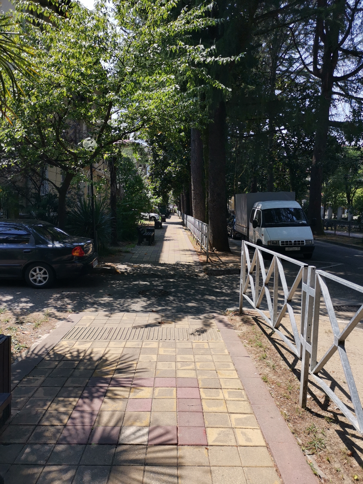 The image shows a sunny day on a sidewalk in Sochi, Russia. The sidewalk is paved with square tiles of beige and reddish-brown colors, bordered by a reddish-brown concrete strip. A metal fence with a zig-zag pattern runs along the right side of the sidewalk, separating it from a road. On the road, a white truck with a gray tarp is parked, and further down, another white vehicle is visible. To the left of the sidewalk, there is a black sedan parked on a dirt area. Lush green trees with dense foliage line the street, casting dappled shadows on the sidewalk and the road. In the background, buildings are partially visible through the trees. The scene appears quiet and typical of a residential or commercial street in Sochi.