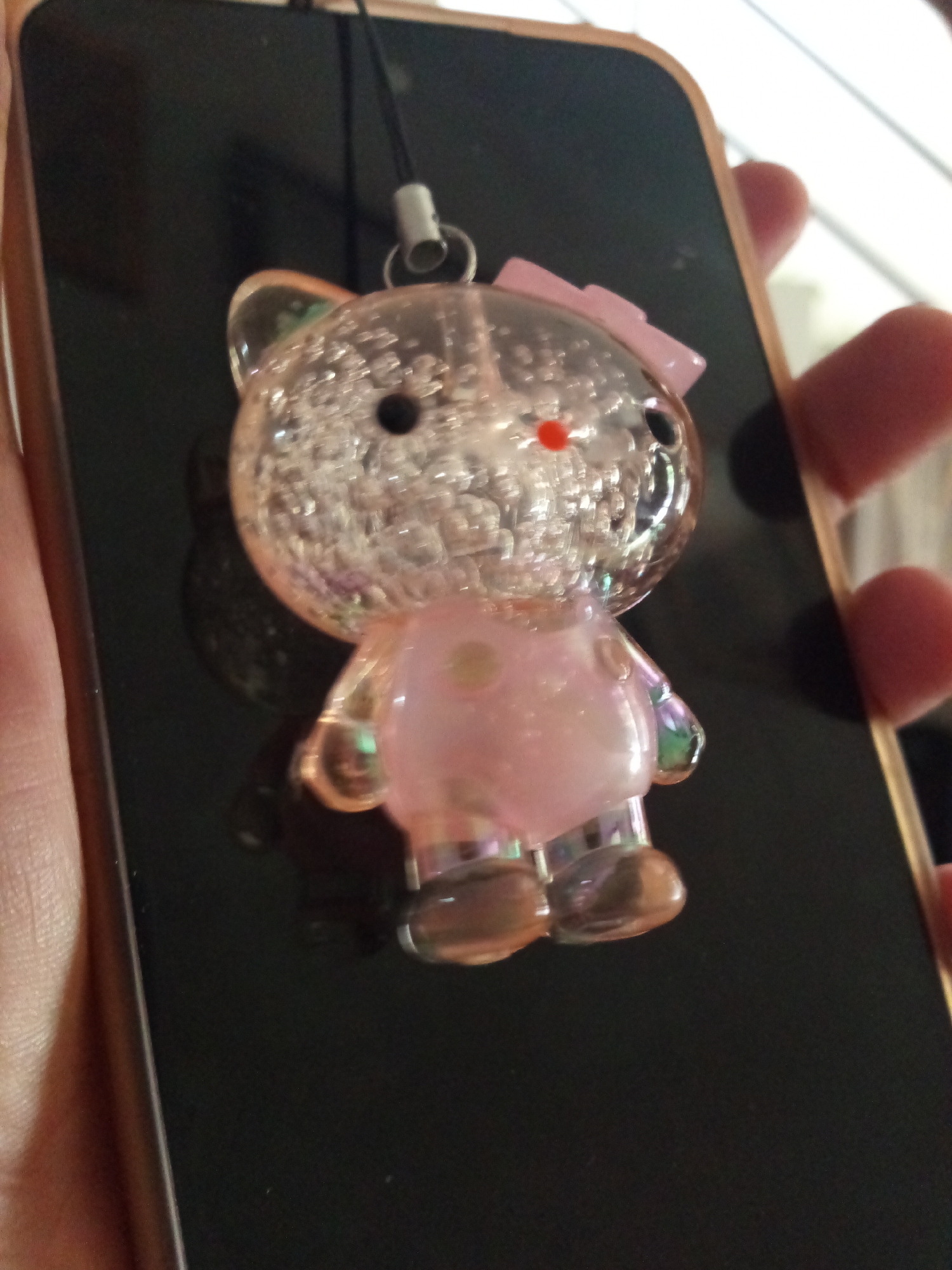 A person holds a black-screened mobile phone with a Hello Kitty charm attached to the top. The charm is translucent, bubbly, and has a pink bow, dress, and features. The phone is presumably in Davao City, Philippines, since that is where the query specifies to locate the scene. However, there is no visual evidence within the image itself to confirm this location.