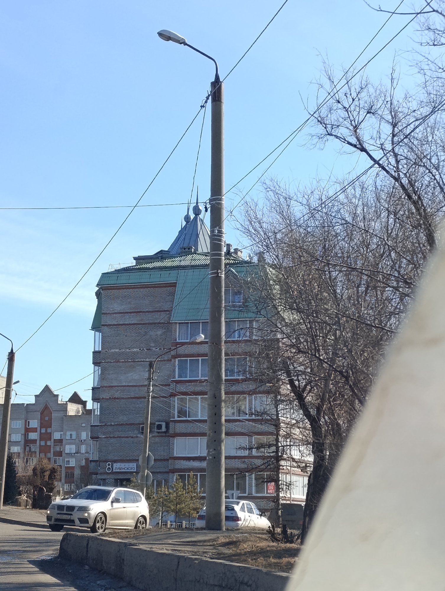 The scene depicts a street view on Dubensky Street 8, Krasnoyarsk, Russia, under a clear blue sky. A prominent multi-story building, constructed with light gray or beige bricks accented by reddish horizontal bands, occupies the mid-ground. It features a distinctive dark green multi-faceted roof with several pointed spires/domes atop its upper section. Rectangular windows with white frames are visible across its facade, along with external air conditioning units.
In the foreground, a gray concrete curb separates an asphalt road from a low, unkempt median of dry grass and dirt. A light-colored BMW X3 SUV (license plate A555 YX 24 RUS) is visible on the street, facing left. Further down and to the right, a white sedan is partially obscured by bare-branched deciduous trees, indicative of late autumn or winter. Multiple utility poles, primarily concrete, stand along the street, supporting numerous overhead power lines that crisscross the sky. One tall concrete pole in the center features a white, oblong streetlamp fixture. A white street sign with a black border is attached to another pole.
To the left of the main building, other multi-story residential or commercial buildings are visible in the background, also featuring brick construction and various roof designs. Bare-branched trees line the street, while a few small evergreen shrubs are positioned near the prominent building and parked vehicles. The lower right portion of the image is partially obscured by a large, out-of-focus, light-colored object, likely part of a vehicle's interior or immediate foreground.