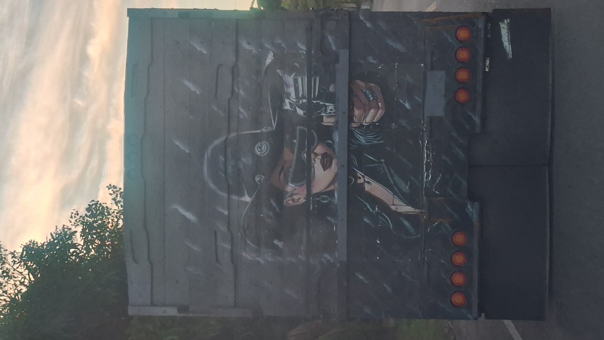 The image shows the rear of a dark-colored truck with a mural painted on it. The mural depicts a woman wearing a cowboy hat and sunglasses, aiming a revolver. She has dark curly hair, bright red lips, and is wearing a black leather jacket and a necklace. The background of the mural appears to be dark and rainy, with streaks of white suggesting falling water. The truck has four orange circular lights arranged horizontally near the bottom, above a dark rectangular section. The truck is on a road, with a tree and a cloudy sky visible in the background above the mural. The location is identified as Lamilieteungoh, Indonesia.