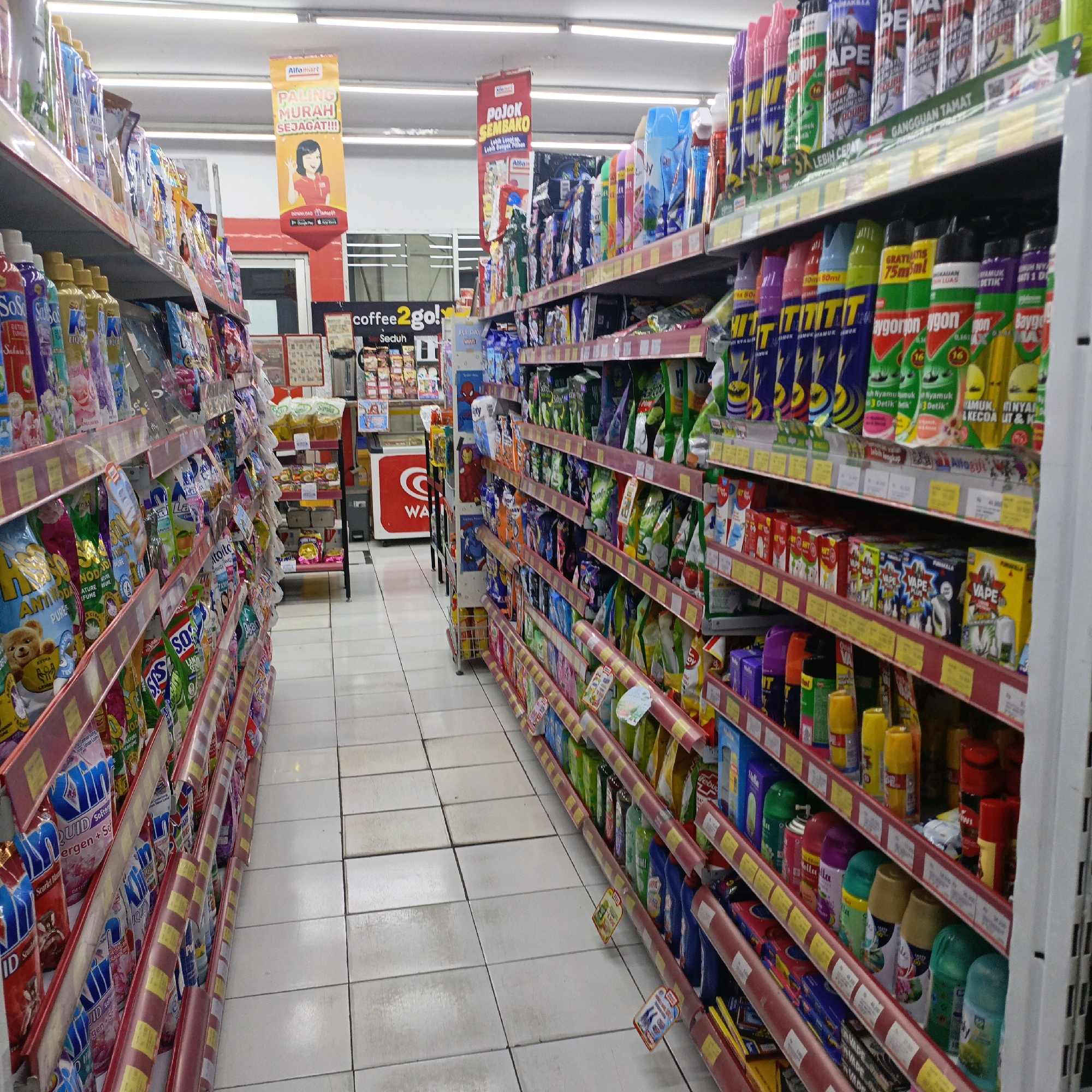 The image shows an aisle in a minimarket, likely Alfamart, in Salatiga, Indonesia. The aisle is stocked with various household products, including detergents, air fresheners, and insect repellents, neatly arranged on shelves. The flooring is tiled in a light color, and fluorescent lights illuminate the scene from above. A red and yellow promotional banner hangs from the ceiling, featuring the Alfamart logo and text in Indonesian. The overall impression is one of a well-organized retail environment, typical of a convenience store in Indonesia.