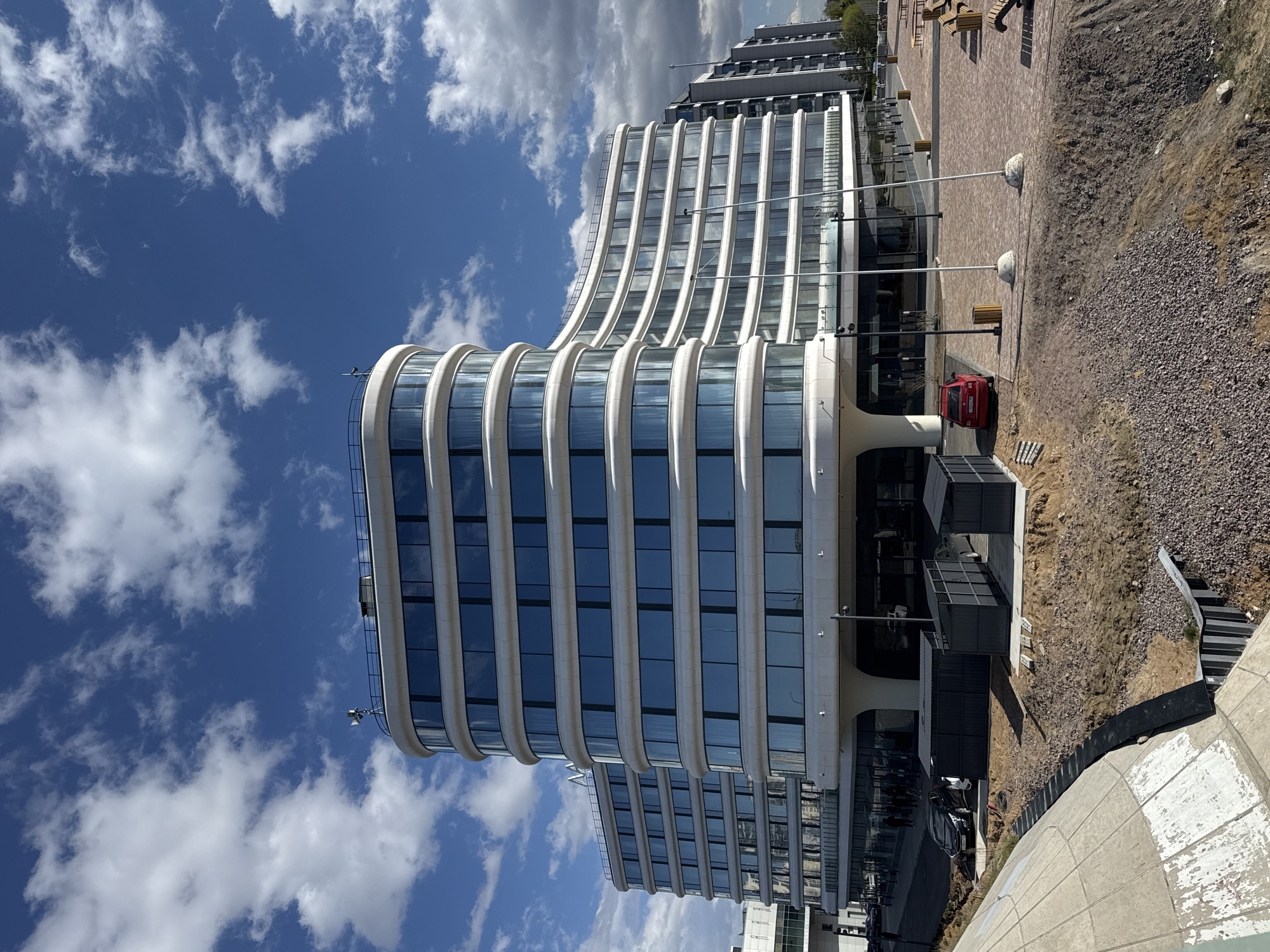 The image depicts a modern architectural building under a blue sky with scattered clouds. The building features a distinctive curved design with numerous windows and a white exterior. A red car is parked near the building. The surrounding environment includes a paved area, landscaping elements, and construction work.
Based on visual clues, this building is likely located in Saint Petersburg, Russia.