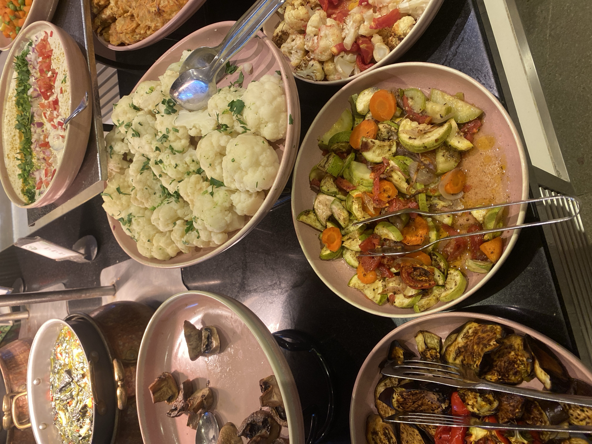 A buffet spread of various dishes is displayed on a dark countertop. In the foreground, a pink oval dish contains grilled eggplant slices with red bell peppers and herbs. Next to it, another pink oval dish holds a mixture of zucchini, carrots, and tomatoes in a light sauce. To the upper right, a pink oval dish is filled with cauliflower florets, some appearing roasted. Further back and to the left, a pink shallow dish holds a creamy dip topped with diced tomatoes, red onions, and chopped herbs. Behind that, a large metal pot contains a green stew with visible ingredients. To the far right, a partial view of a dish with fried food is visible. Serving utensils, including forks and spoons, are present in several of the dishes. The lighting suggests an indoor setting, possibly a restaurant or food service establishment in Aksu, Turkey, given the context of the prompt. There are no people visible in the image.