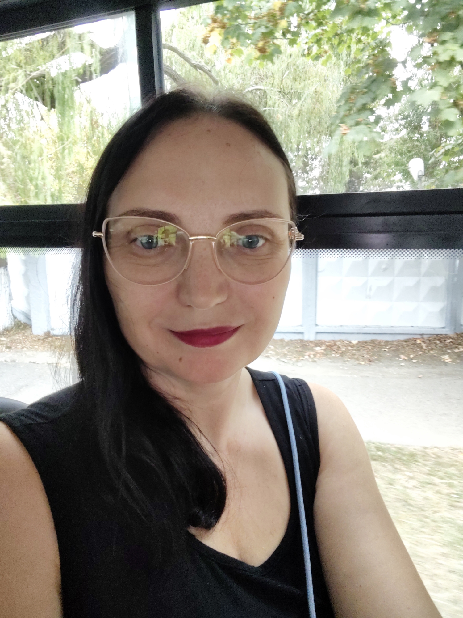 A woman with dark hair and glasses is taking a selfie. She is wearing a black tank top and dark lipstick. She is sitting in what appears to be a vehicle, possibly a bus, with a window to her left showing trees and foliage. The background behind her is a patterned wall and some foliage. The image is taken from the chest up, focusing on her face. No specific location in Sochi, Russia can be determined from the visual information provided.