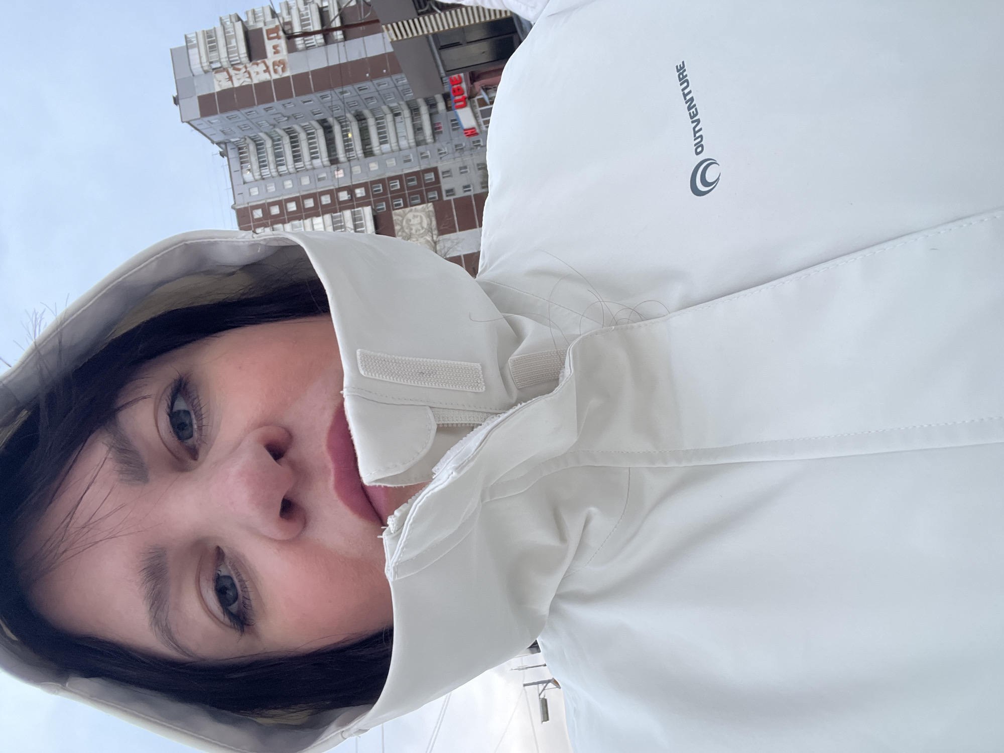 A selfie captures a person outdoors, framed by a light-colored, possibly white or off-white, hooded jacket. The image, originally rotated 90 degrees clockwise, shows the person looking directly at the camera. They have dark hair and light-colored eyes (blue/grey). The hood of the jacket is up, framing their face, and the jacket features visible Velcro fasteners around the neck/collar area.
On the right shoulder area of the jacket, the brand name "OUTVENTURE" is visible, accompanied by a circular logo that resembles a stylized crescent moon or 'C' shape.
In the background, a large, multi-story building with numerous windows dominates the urban skyline. The building appears to have sections of light and brown cladding. Above the building, the sky is overcast and pale grey, indicating daytime. Bare tree branches are faintly visible on the left side, suggesting cooler weather, possibly winter or early spring.
Notable Cyrillic text is visible on the building in the background, including "ТЦ" (likely an abbreviation for "Торговый Центр" or Shopping Center), consistent with the provided location context of Perm, Russia. The overall impression is a candid moment captured in an urban setting during an overcast day.